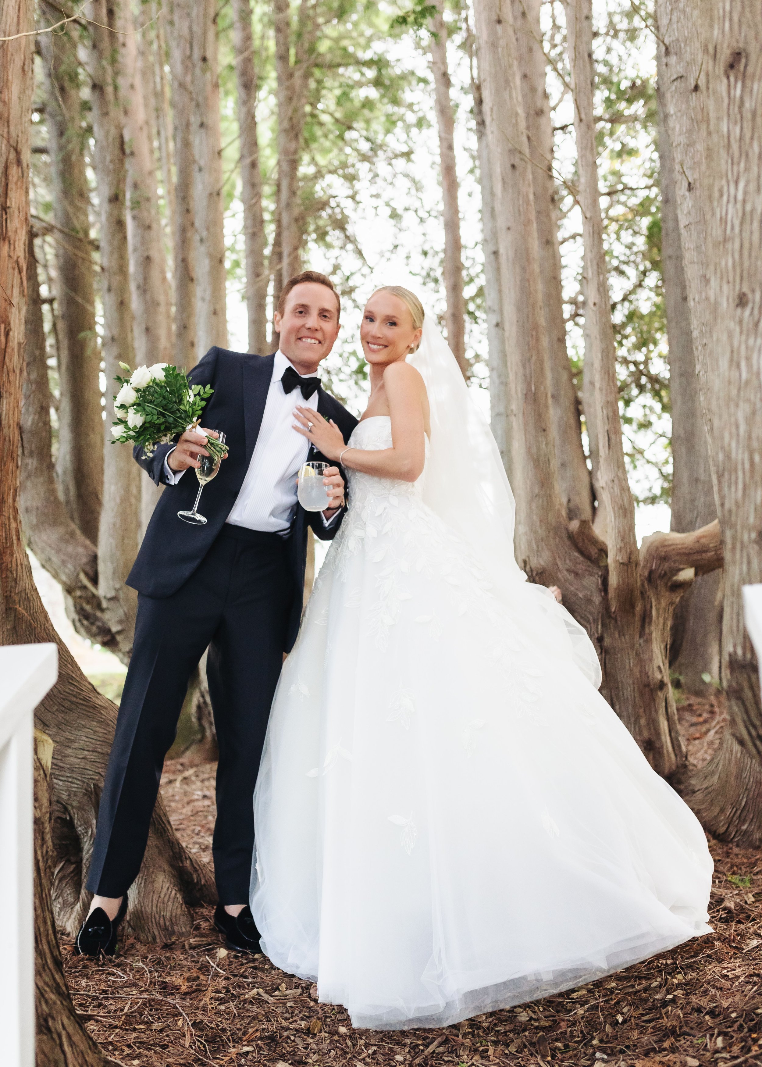 A newly married couple smiling and posing in a wooded area with tall trees, the bride wearing a white wedding gown and veil, and the groom in a black tuxedo holding a bouquet and a glass of drink.