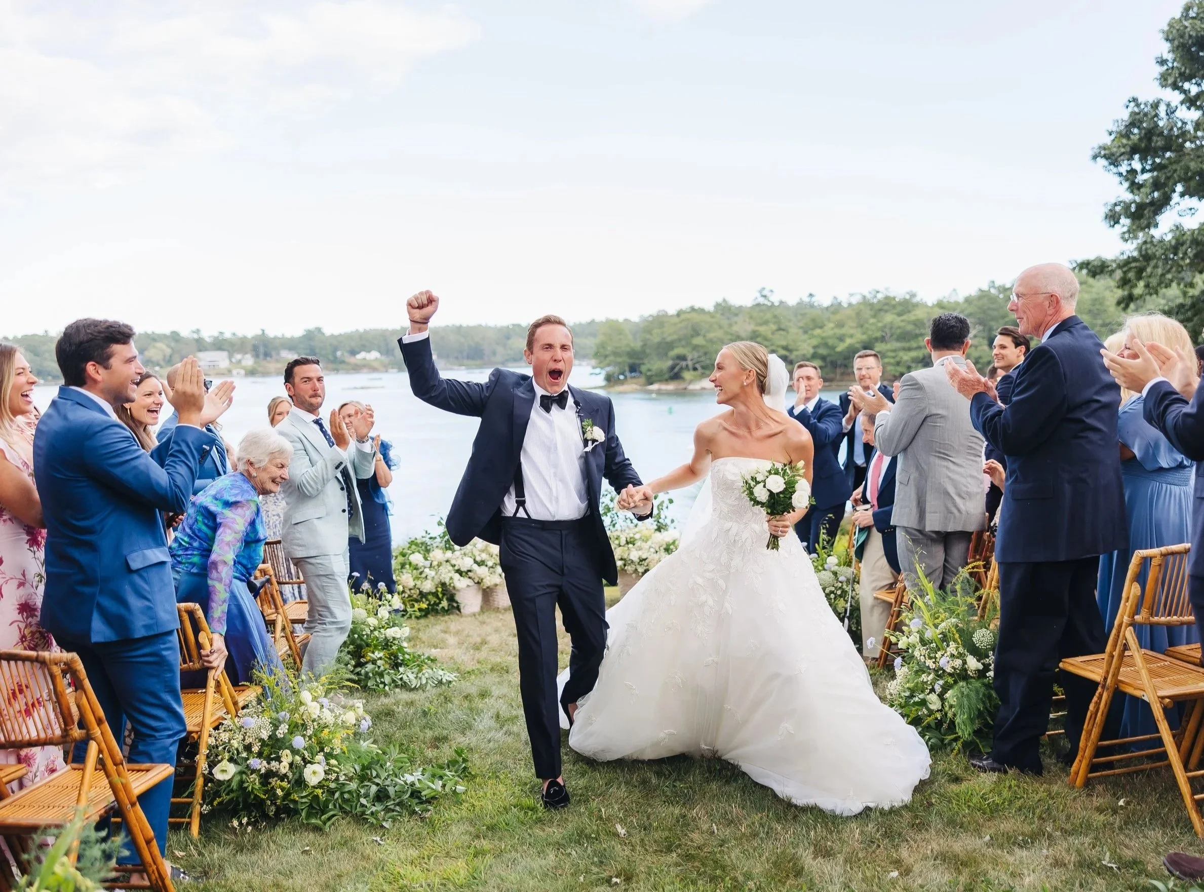 A newlywed couple is walking down the aisle of an outdoor wedding ceremony. The groom is wearing a dark tuxedo with a bow tie, raising his arm in celebration, and the bride is holding a bouquet of white flowers, smiling. Guests are clapping and cheering, and the ceremony is taking place by a body of water with trees in the background.