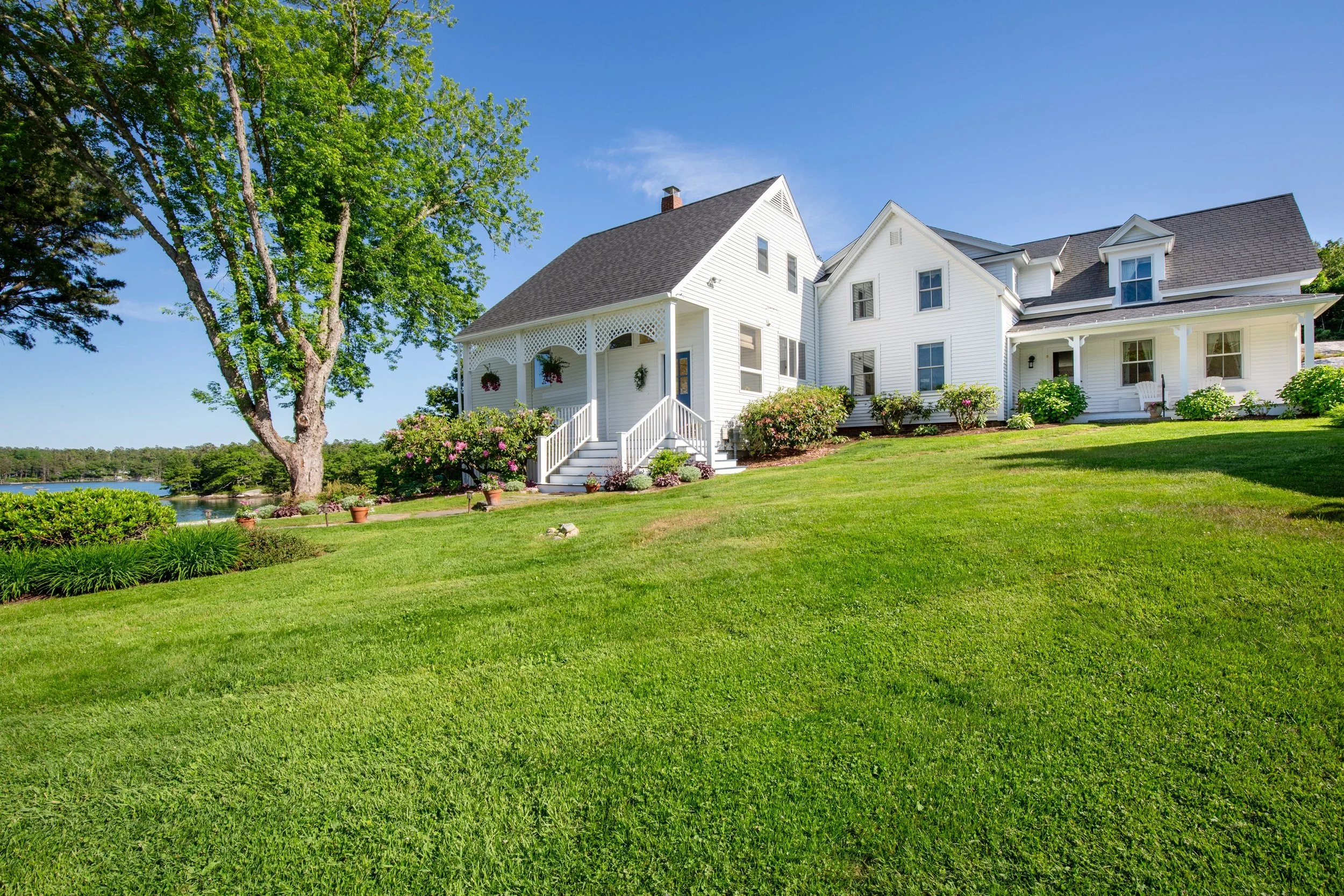 A white, two-story house with a porch, surrounded by a well-maintained green lawn and bushes, with a large tree nearby and a body of water in the background under a bright blue sky.