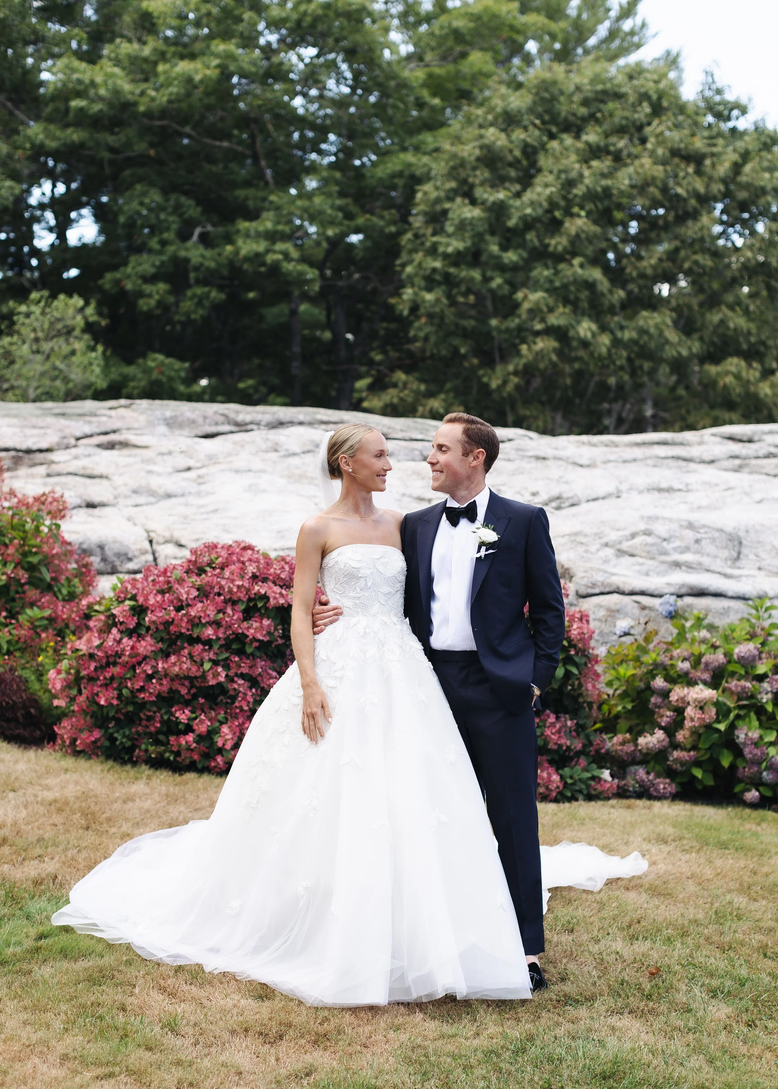 A bride in a white wedding gown and a groom in a black tuxedo with a bow tie are standing on grass in front of pink flowers and green trees, smiling at each other.