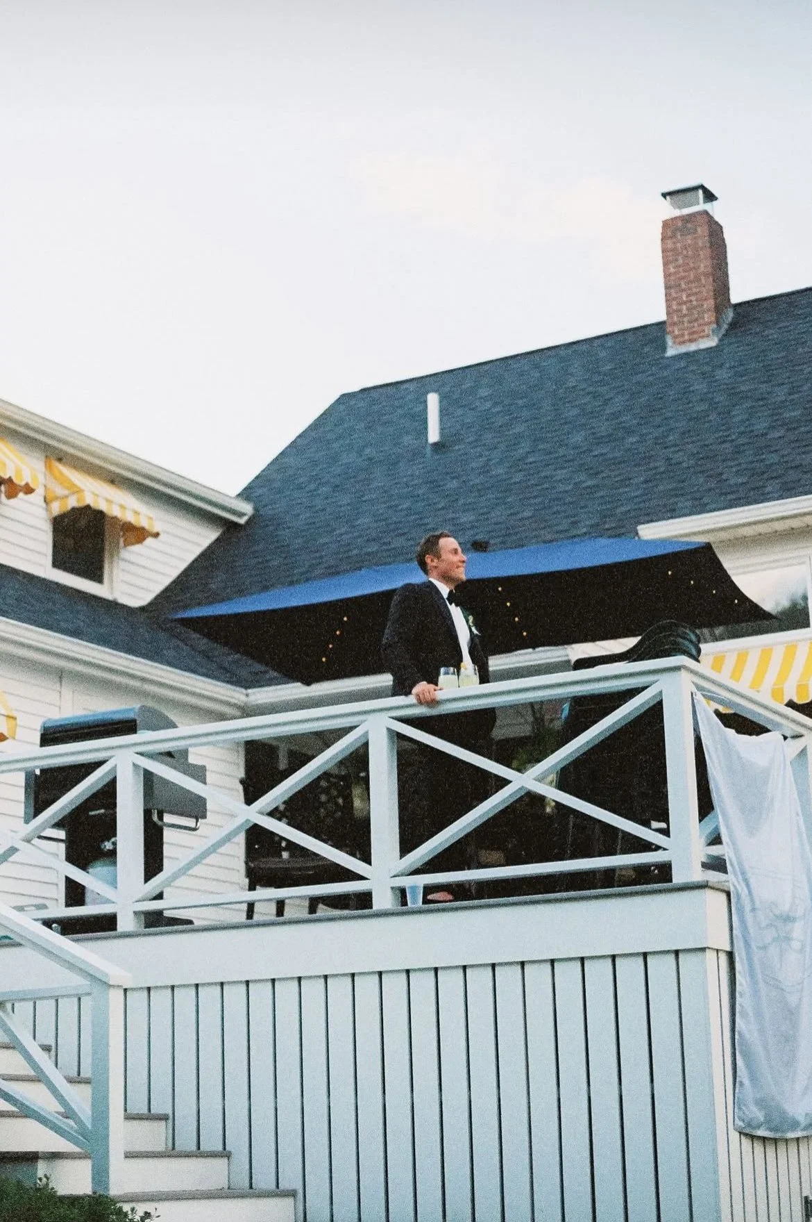 A man in a tuxedo standing on a house's second-story balcony decorated with string lights.
