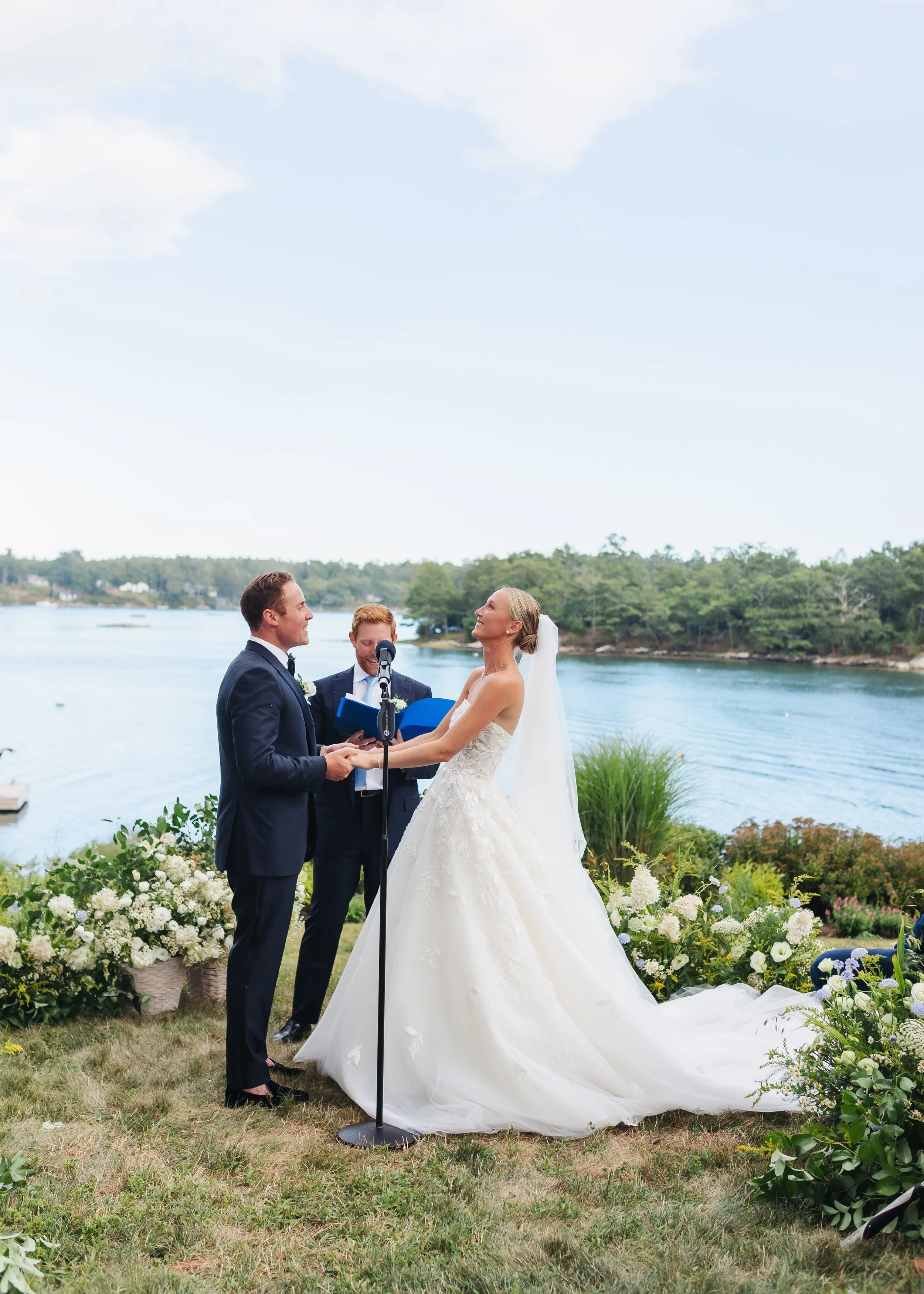 A couple is getting married outdoors by a river, with an officiant present, and the bride in a white wedding dress and veil.