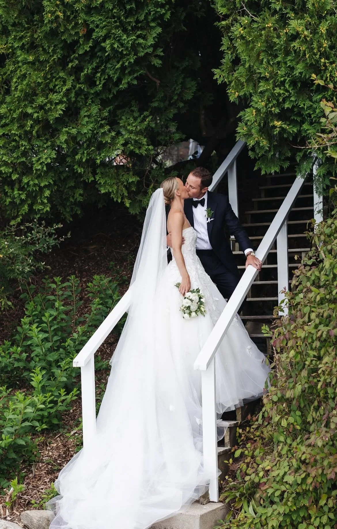 A bride and groom sharing a kiss on a staircase outdoors, surrounded by greenery, with the bride holding a bouquet of white flowers.