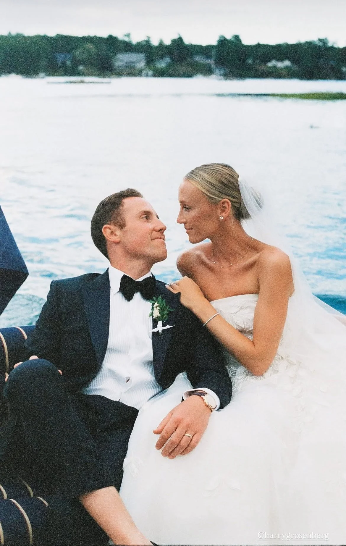 A bride and groom in wedding attire on a boat, with the bride touching the groom's shoulder, on a body of water with trees and buildings in the background.