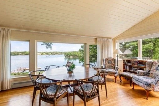 Living room with large windows overlooking a body of water, dining table with six chairs, and vintage armchairs with a side table and lamp.