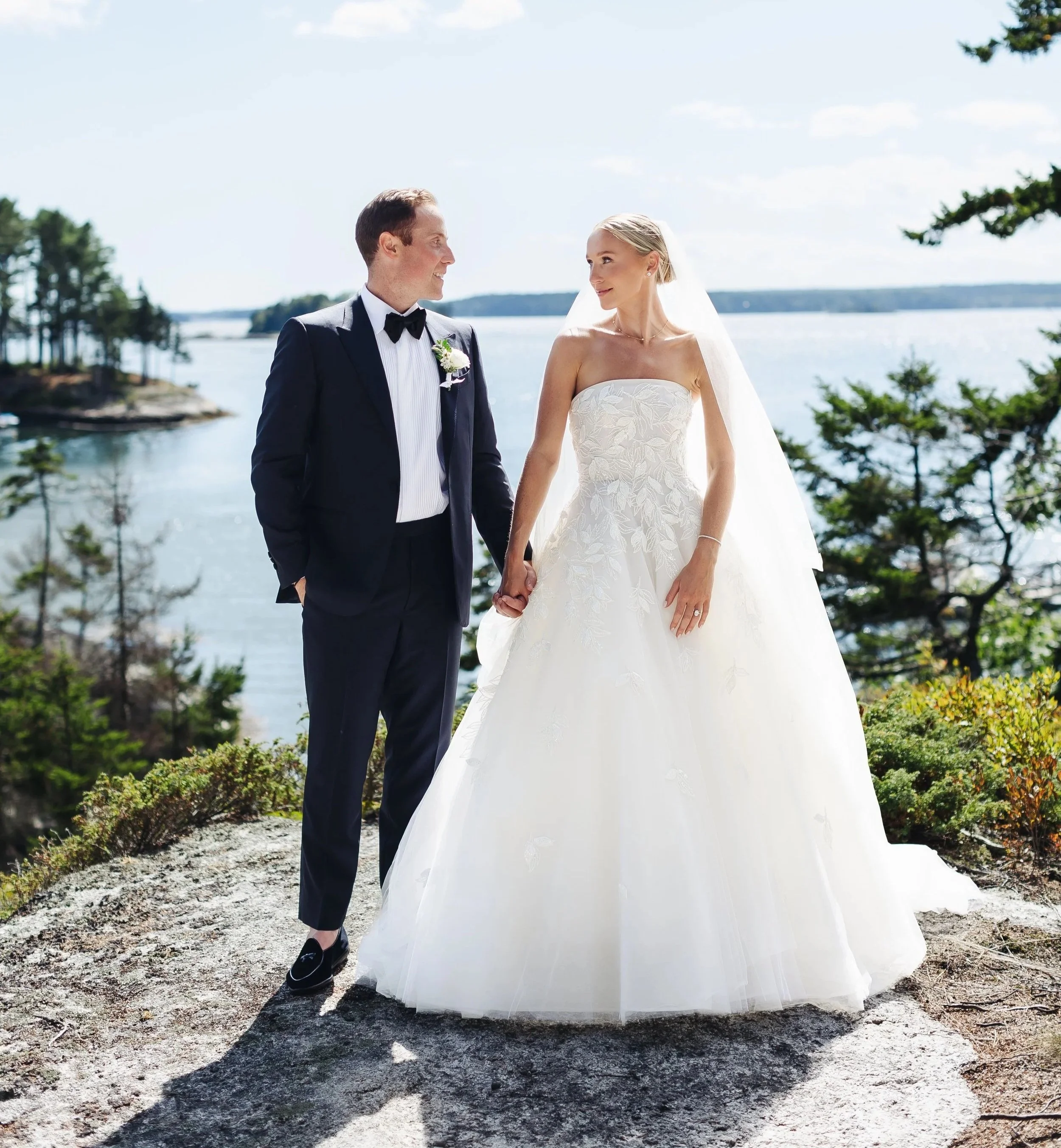 A bride and groom holding hands outdoors near water with trees in the background, the groom wearing a black tuxedo and bow tie, and the bride in a strapless floral wedding gown with a veil.