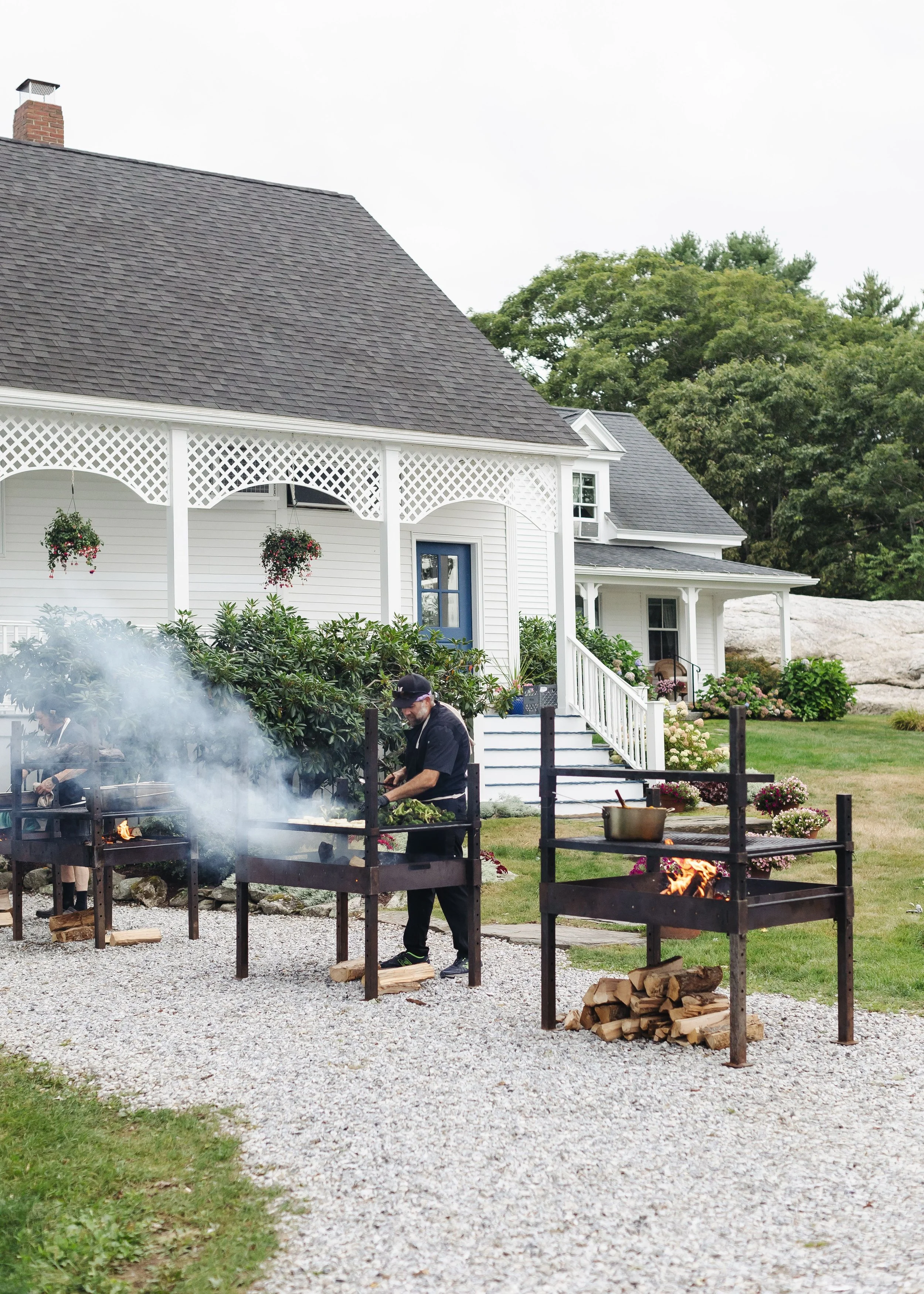 A man wearing a black cap and jacket standing next to a row of outdoor fire pits, with logs nearby, in front of a white house with decorative trim and greenery, under a cloudy sky.