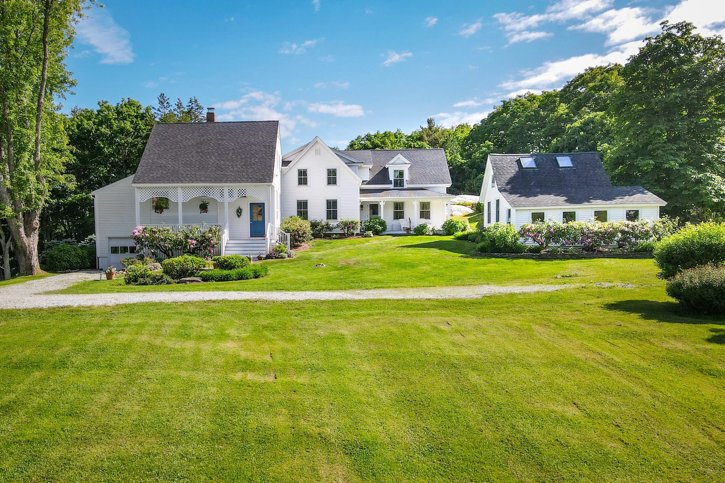 A white house with a gray shingled roof, surrounded by green trees and a well-maintained lawn. The house has a front porch with hanging plants and flower beds, with a clear blue sky above.