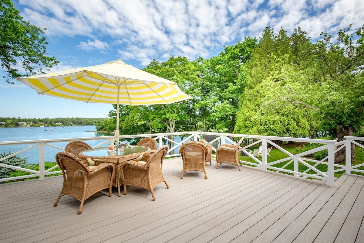 A spacious outdoor deck with wicker chairs and a glass-topped table beneath a yellow and white striped patio umbrella, overlooking a harbor with lush green trees and a partly cloudy sky.