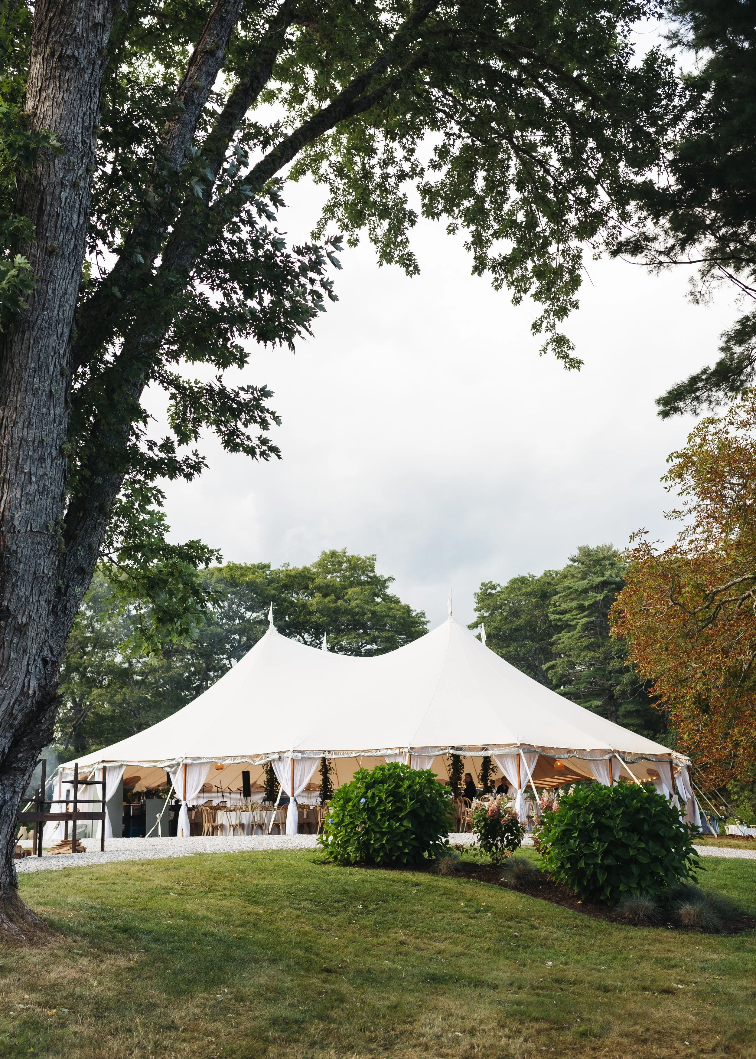 Large white event tent set up outdoors in a lush green garden with mature trees surrounding it, and decorated with flowers at the entrance.
