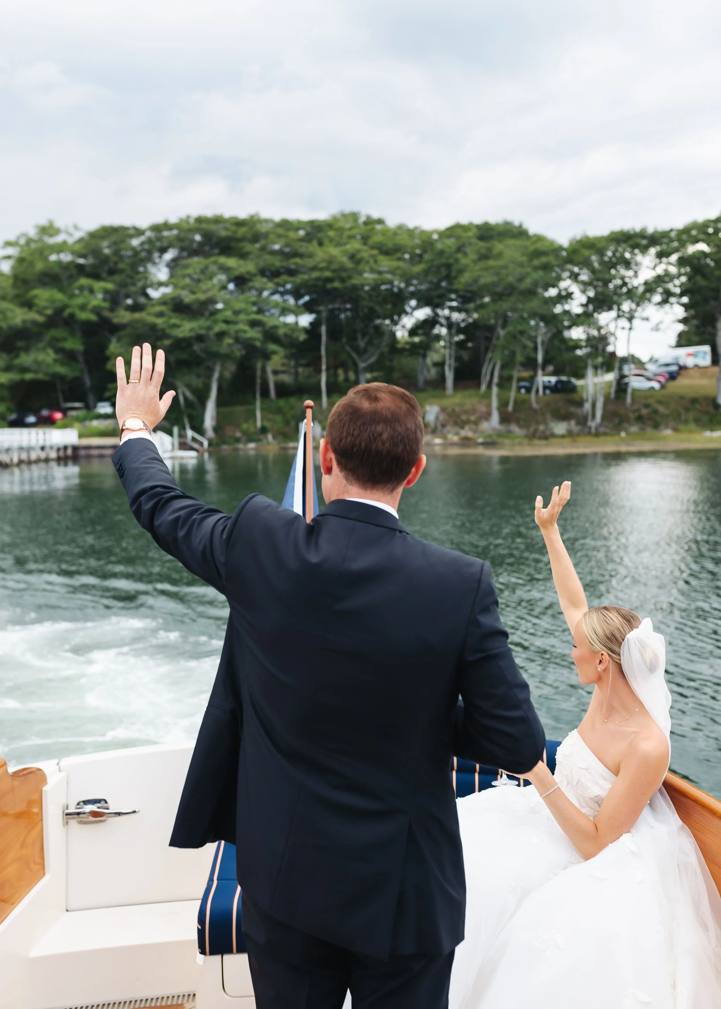 A bride and groom on a boat, with the groom in a black suit and the bride in a white wedding dress, waving to people on land during their wedding celebration.