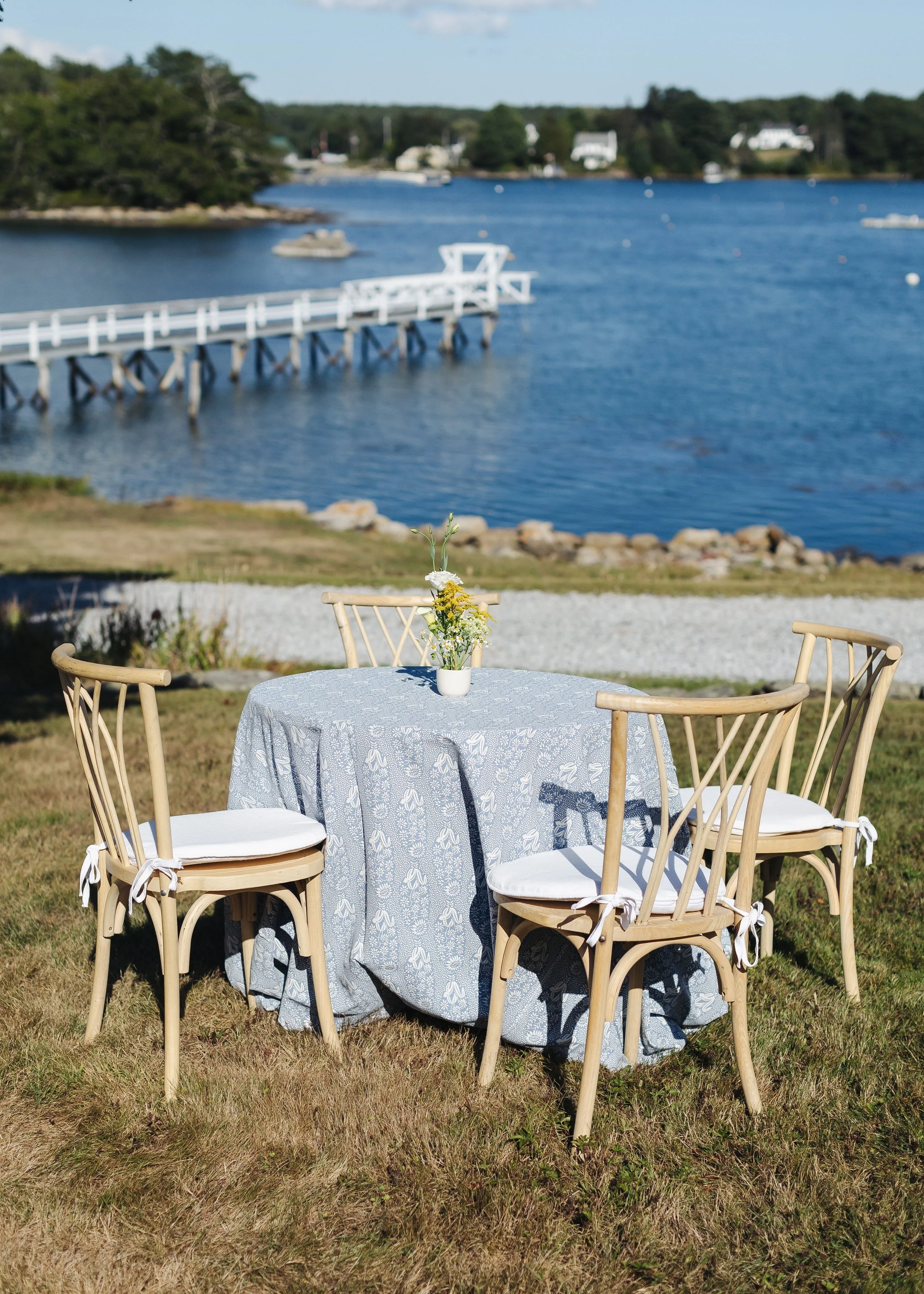 Outdoor table setup with four chairs and a small flower arrangement, overlooking a waterway with boats and a white dock in the background.