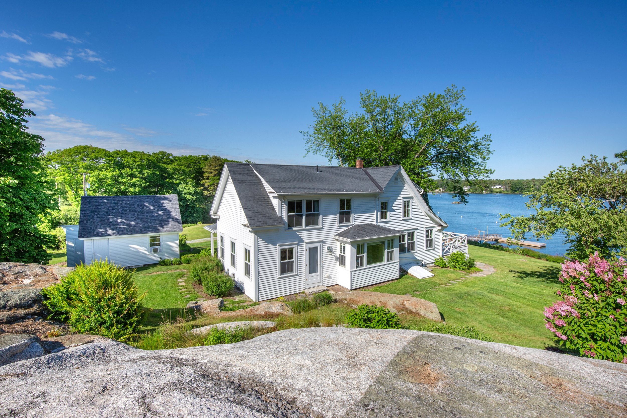 A white house by a body of water, with green trees and a clear blue sky.