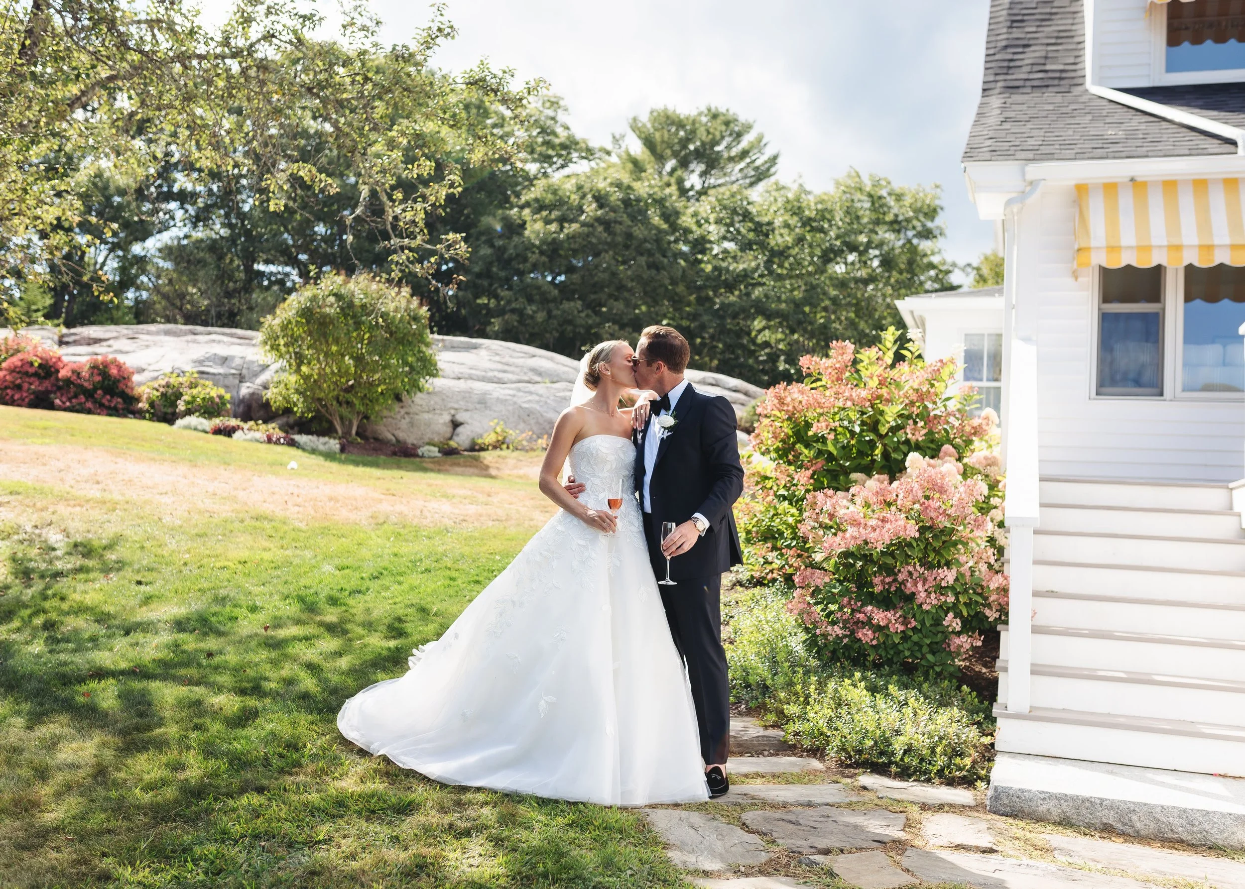 A newlywed couple sharing a kiss outdoors on a sunny day, with the bride in a white wedding gown holding a glass of champagne, and the groom in a tuxedo, standing next to a white house with a garden of pink flowers.