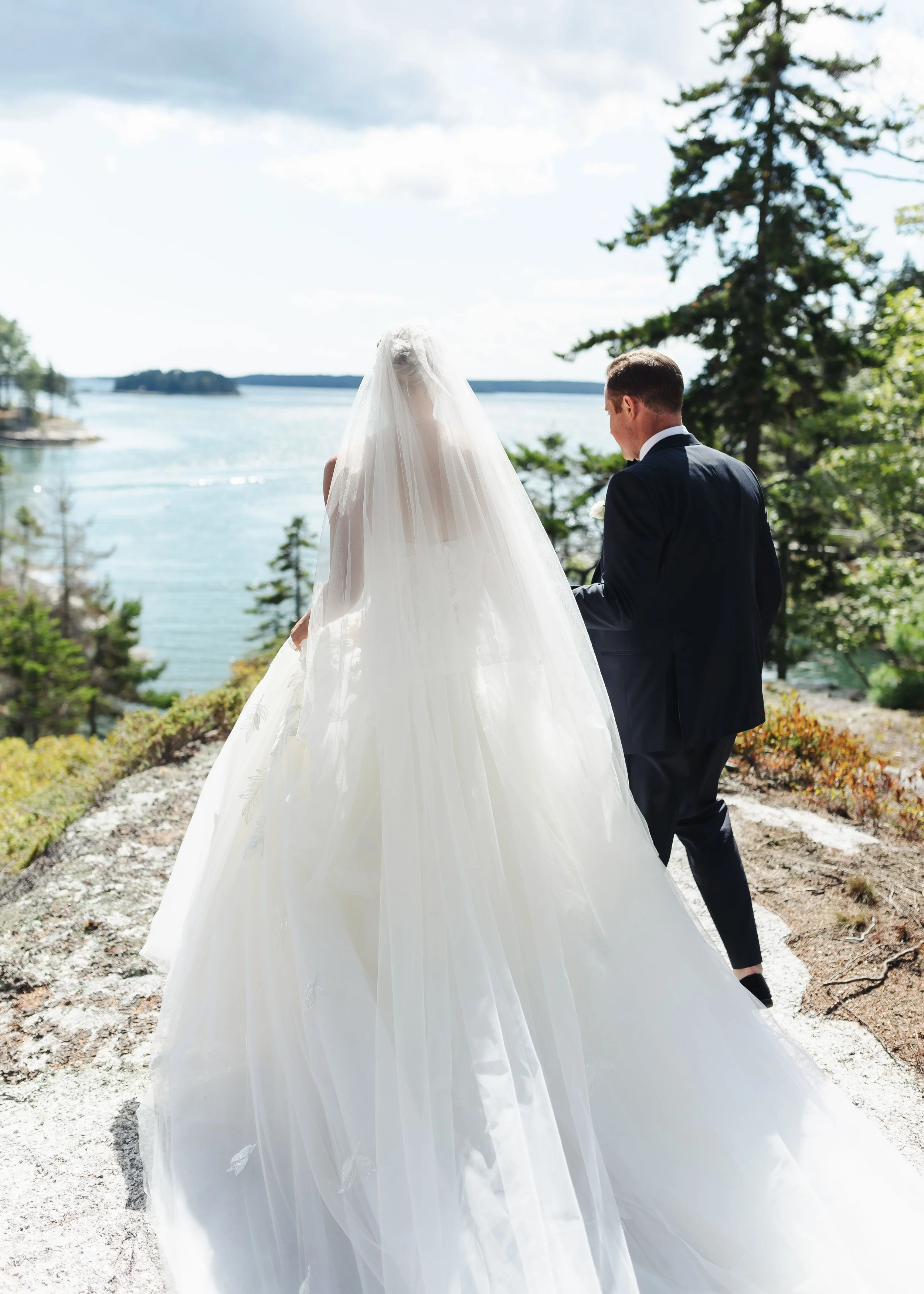 A bride and groom standing outdoors near a body of water, with trees and islands in the background, captured from behind during a wedding ceremony.