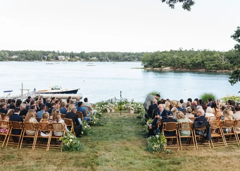 An outdoor wedding ceremony by a river with guests seated on wooden chairs, decorated with flowers, facing the water with boats and trees in the background.