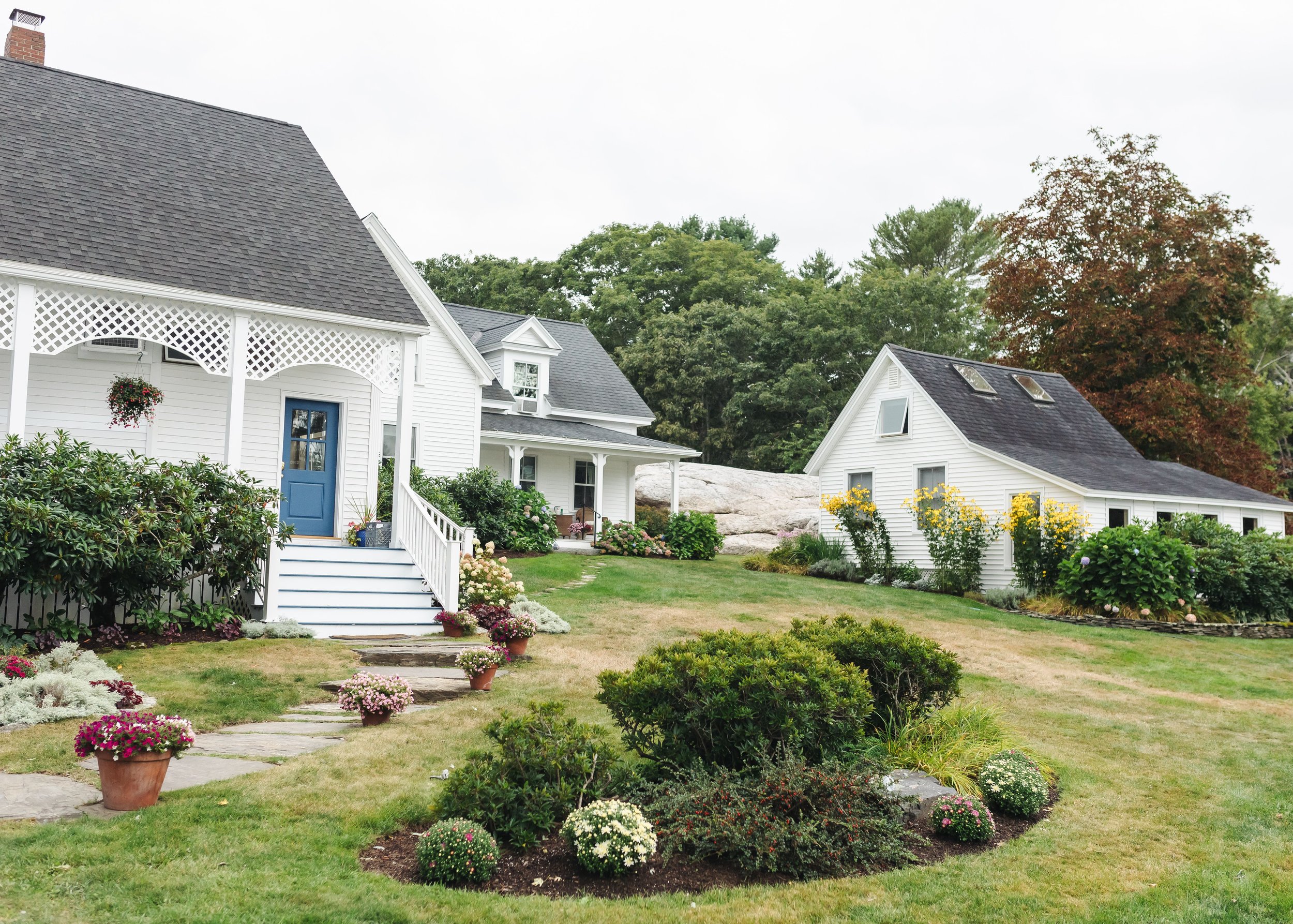 A white house with a blue door and a porch with lattice trim. The front yard has various flowering plants and shrubs, and a stone pathway lined with potted flowers. There are trees in the background, and a second house with skylights on the roof is v