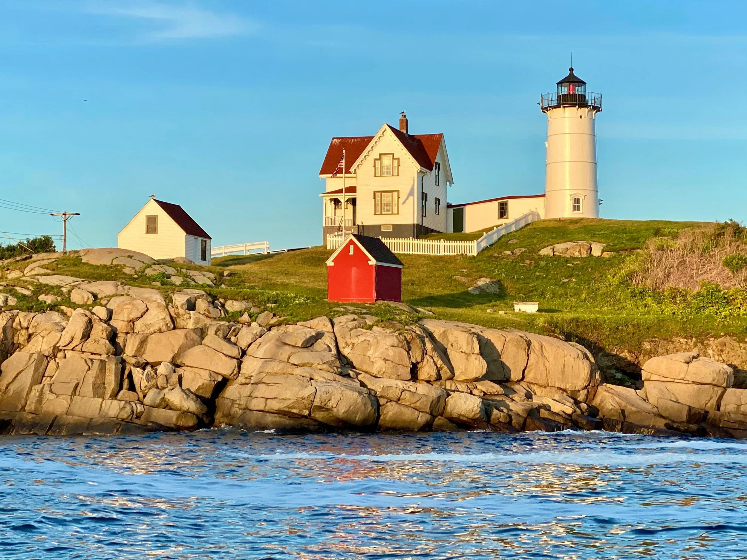 A lighthouse and several houses standing on a grassy hill near rocky shoreline, with water in the foreground and a blue sky above.