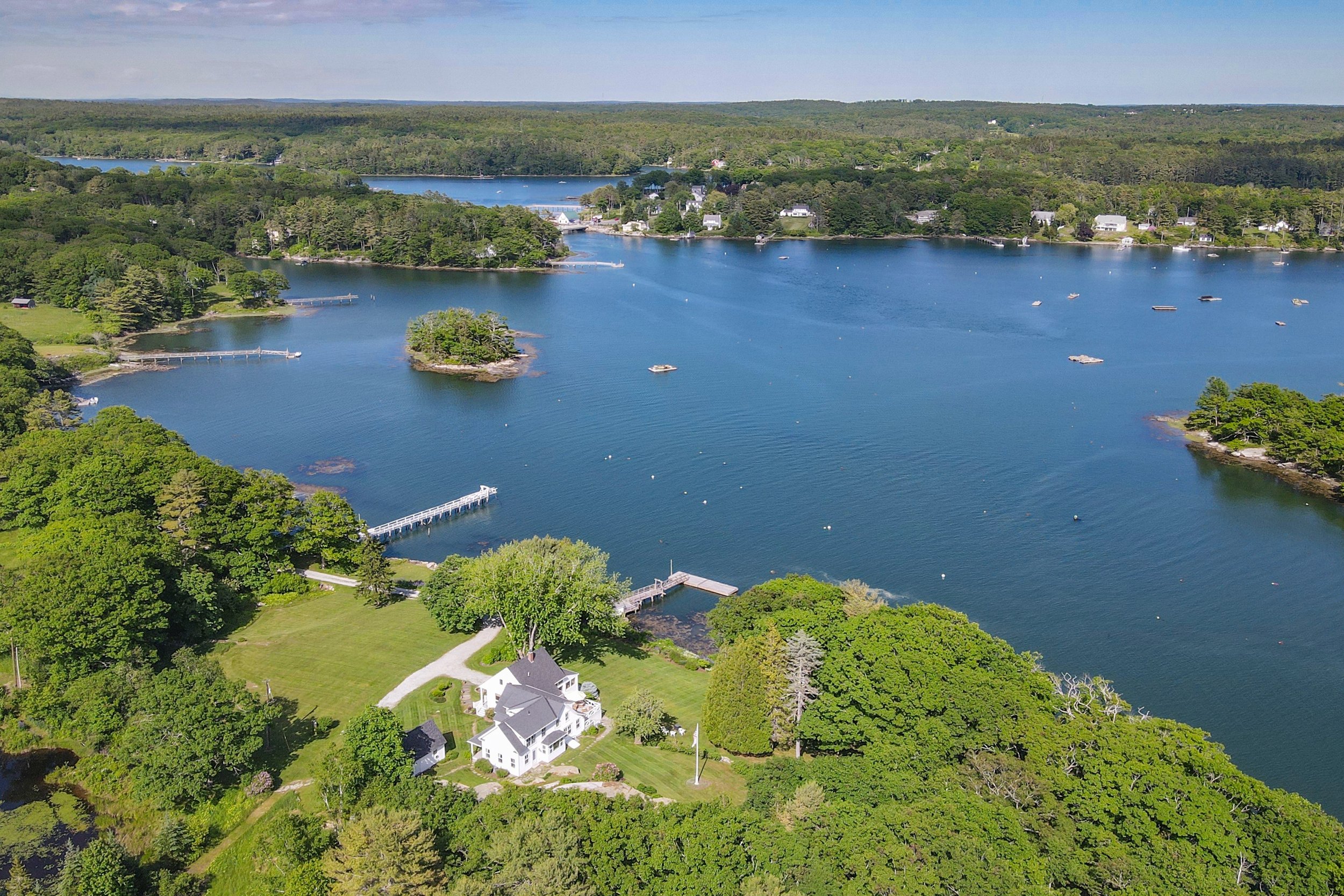 Aerial view of a harbor with a house the shoreline, surrounded by lush green trees, with a small island in the water and multiple docks extending into the water.