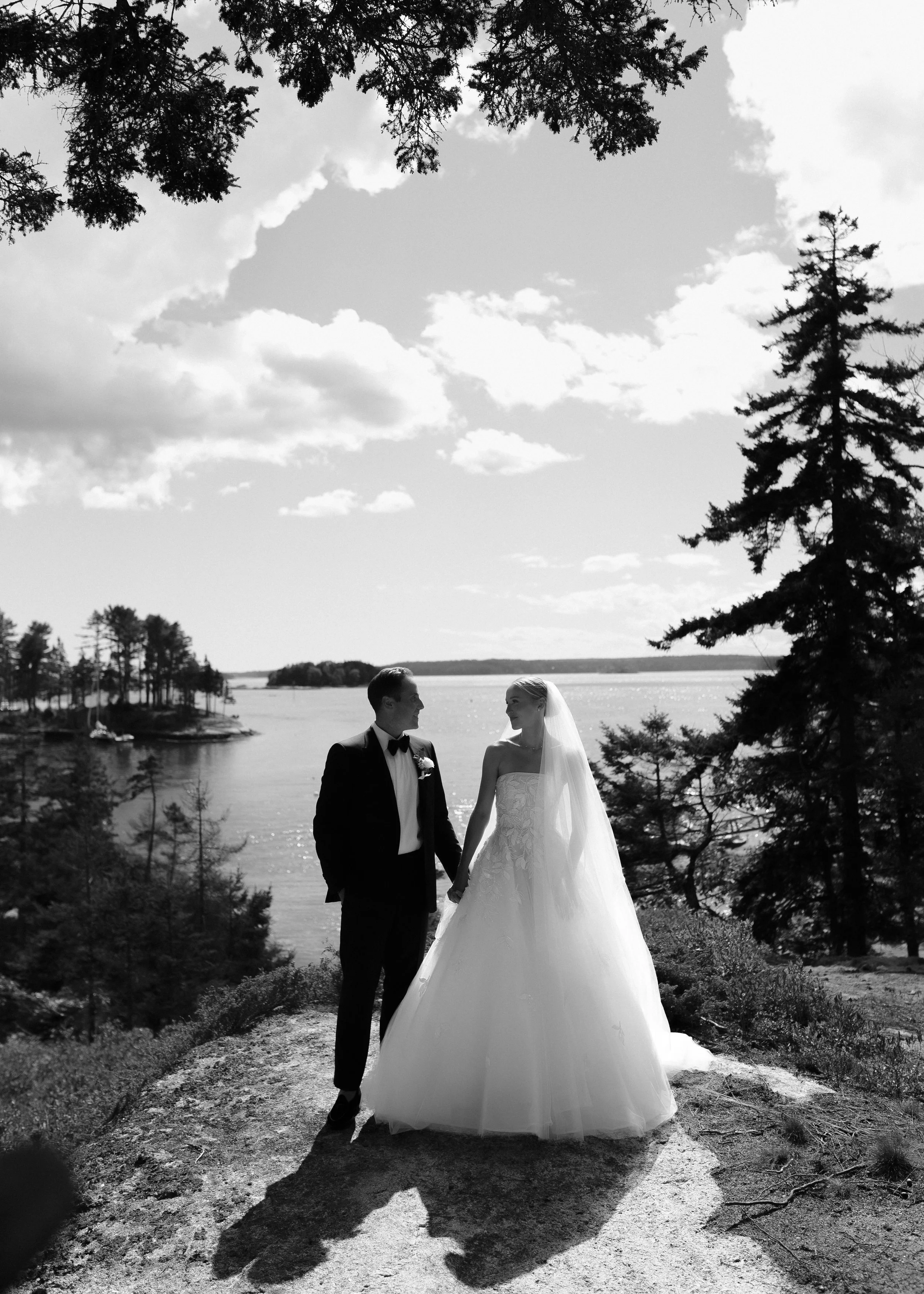 A black and white photo of a bride and groom holding hands, standing outdoors near a body of water, with trees and a partly cloudy sky in the background.