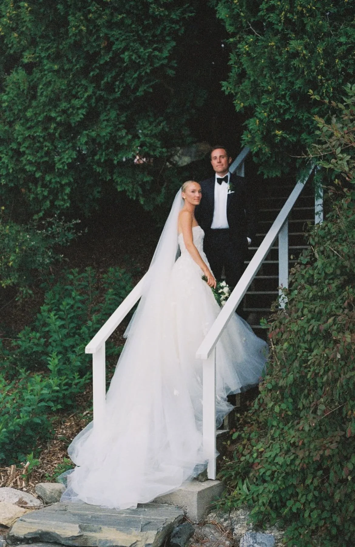 Bride and groom standing on an outdoor staircase surrounded by greenery.