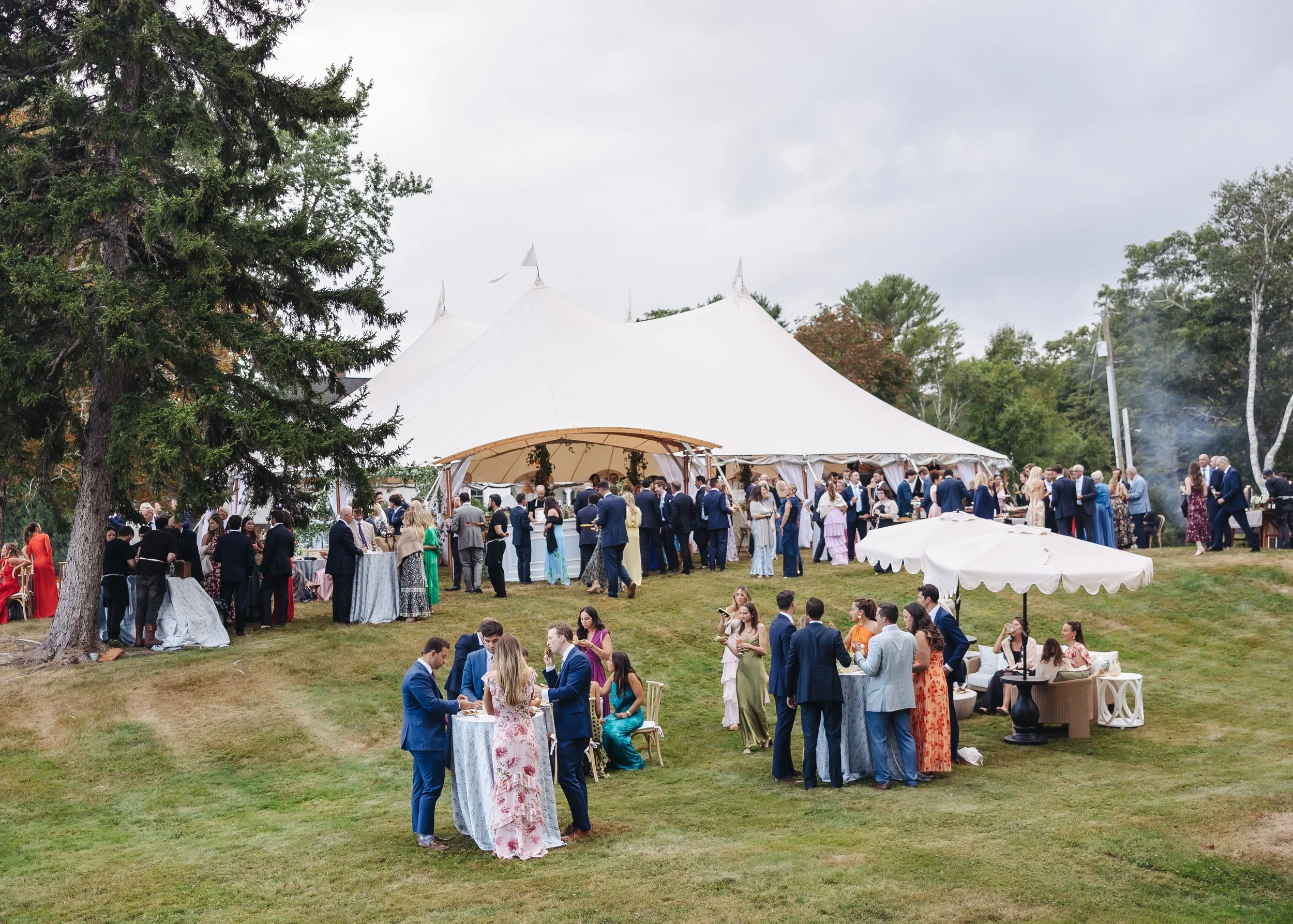 Outdoor wedding reception with guests dressed in formal attire, under a large white tent, surrounded by trees and greenery.