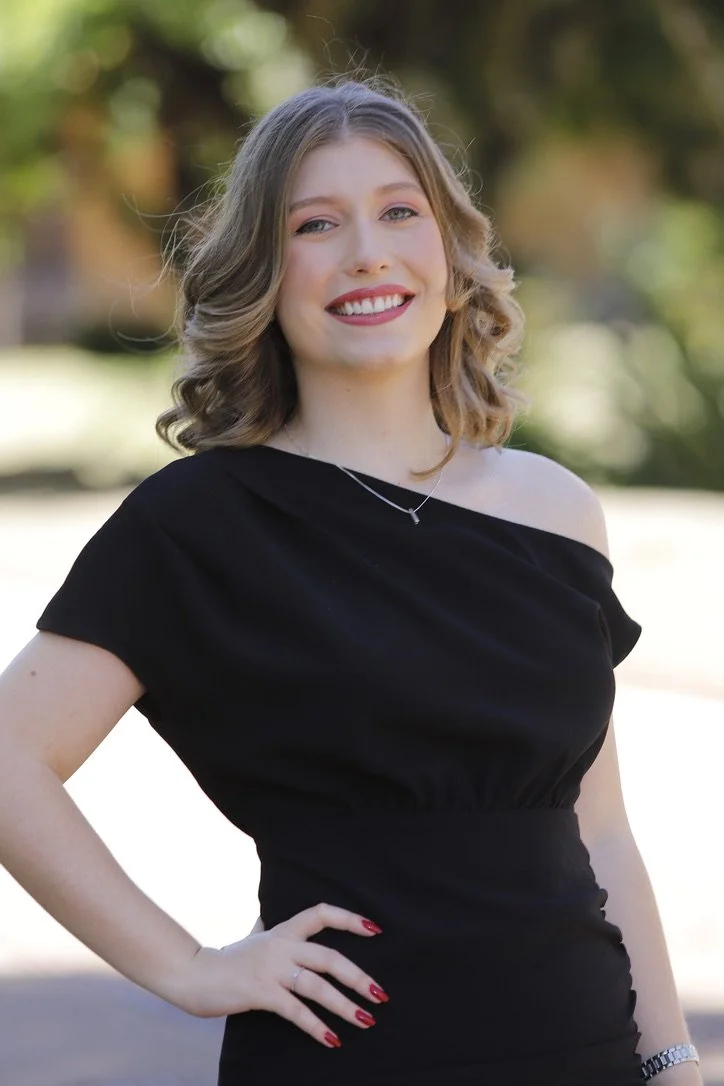 Mulher sorridente com cabelo loiro cacheado usando vestido preto e acessório de colar, ao ar livre com fundo de árvores.