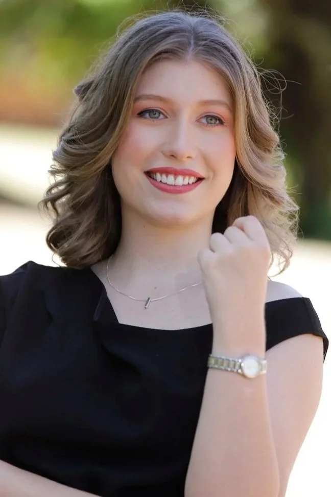 Mulher com cabelo loiro, olhos azuis, sorrindo, fazendo um gesto de força com o punho fechado, usando vestido preto, colar e relógio