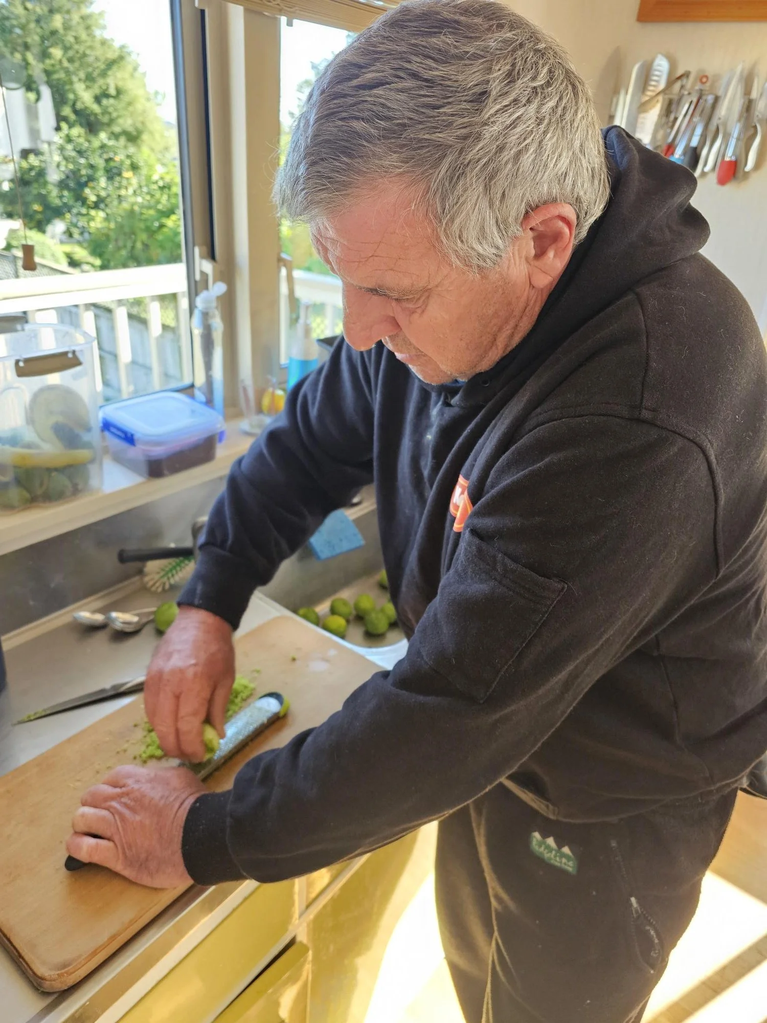 An elderly man with gray hair wearing a black hoodie is chopping green vegetables on a wooden cutting board in a kitchen with sunlight coming through the window. There are kitchen utensils hanging on the wall, and containers and kitchen supplies on the counter in the background.