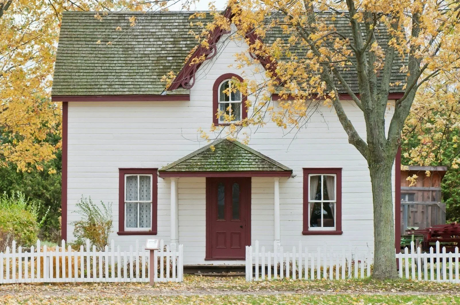 A two-story white house with red trim, a small front porch with columns, and a dark red front door, surrounded by trees with yellow autumn leaves and a white picket fence.