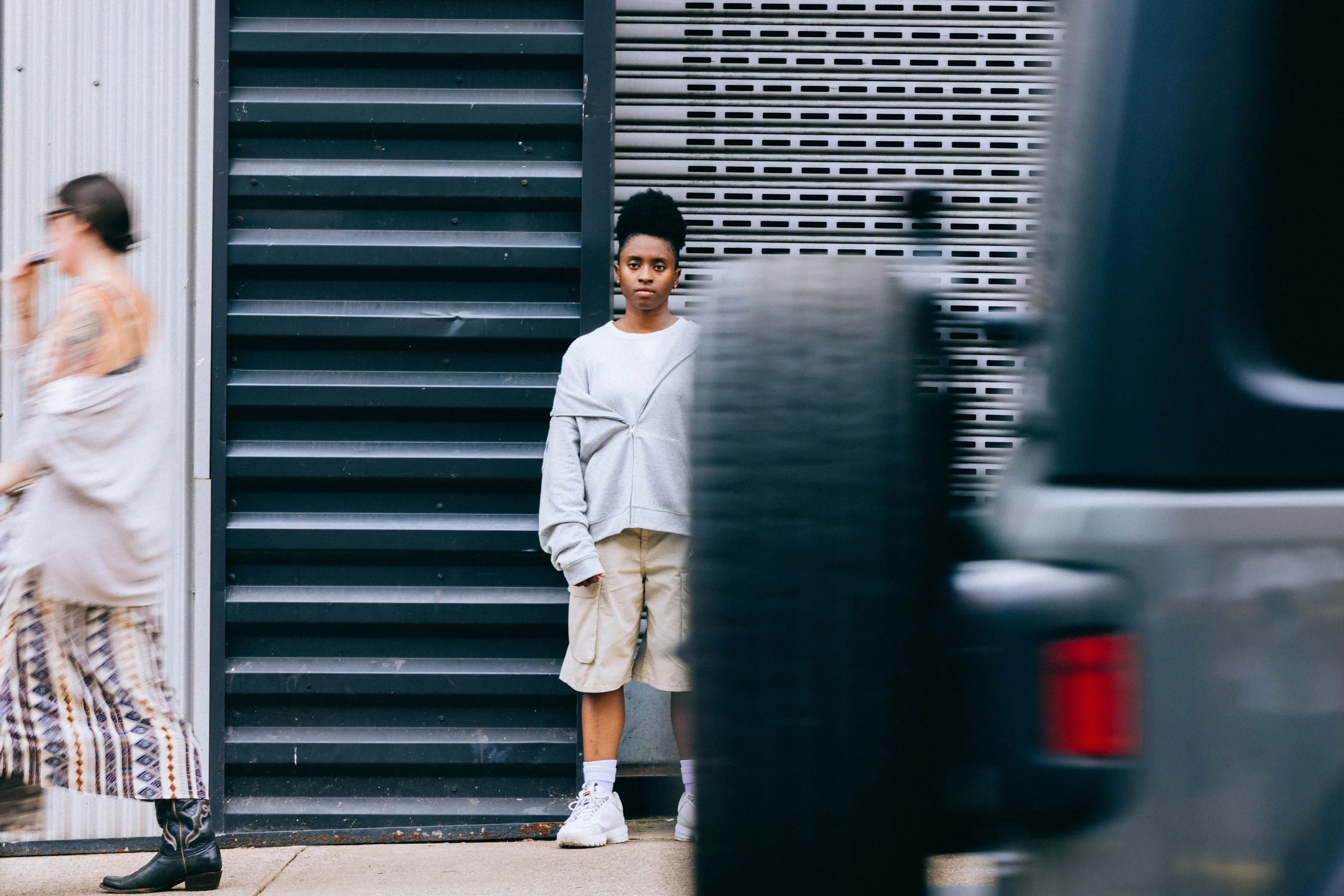 A young woman standing against a modern, industrial-style metal wall, looking directly at the camera. She's wearing a light-colored sweatshirt, beige shorts, white socks, and white sneakers. A woman in a patterned dress and black boots is walking by in the foreground, and part of a car is visible on the right side of the image.