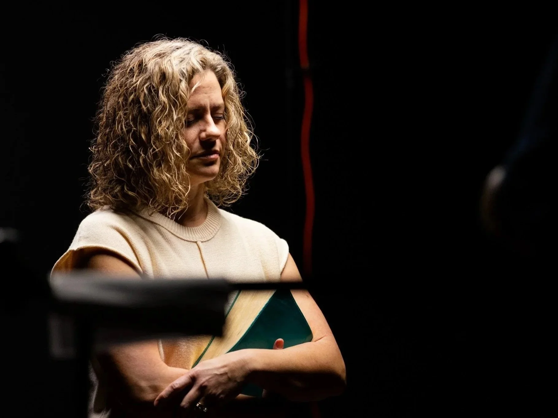 A woman with curly blonde hair is standing with her eyes closed, holding a book or folder in front of her, against a dark background with red and black elements.