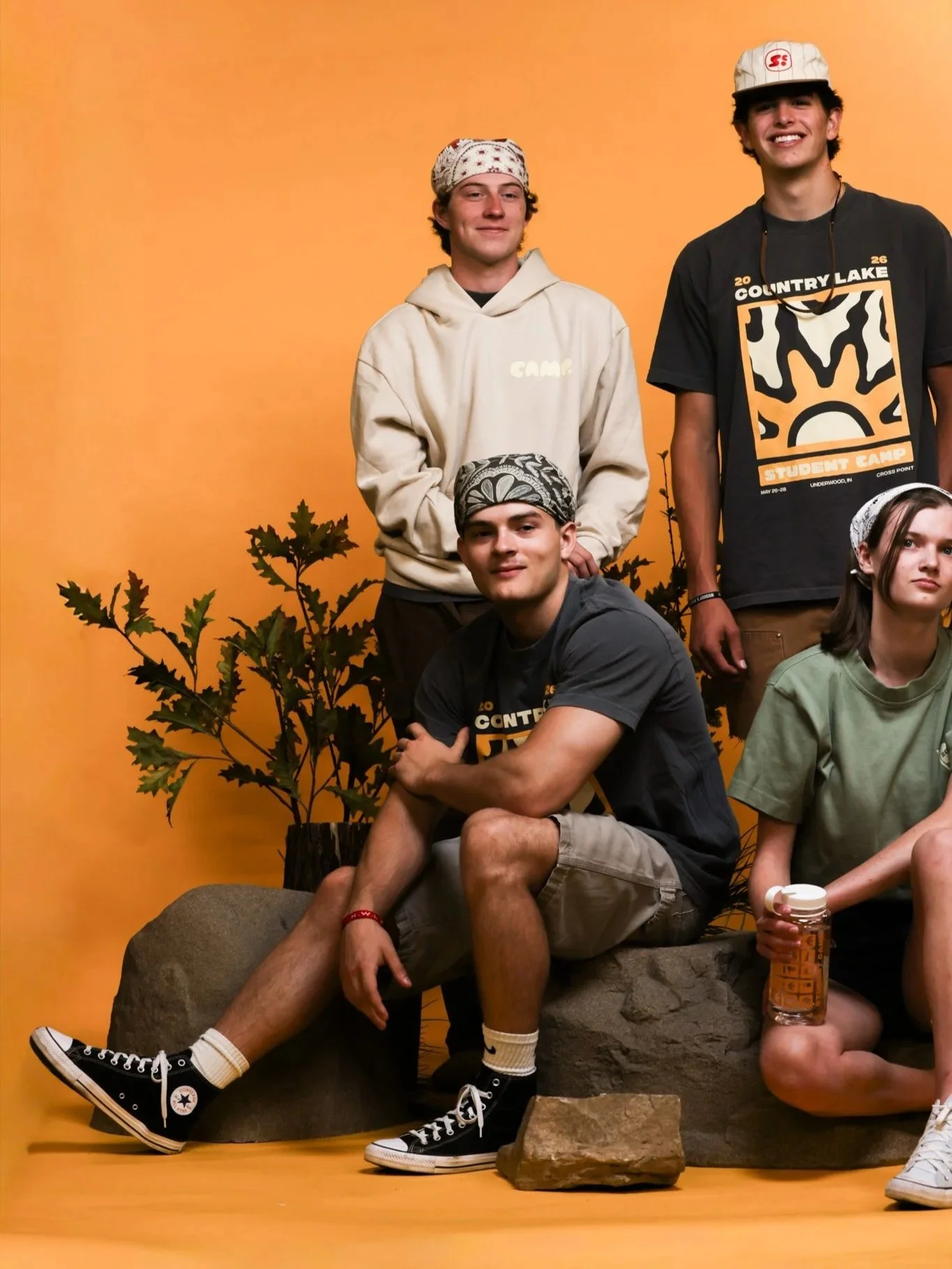 Group of five young people with casual clothing and headwear, posing in front of an orange background with a potted plant, some seated on a large rock and others standing, smiling.