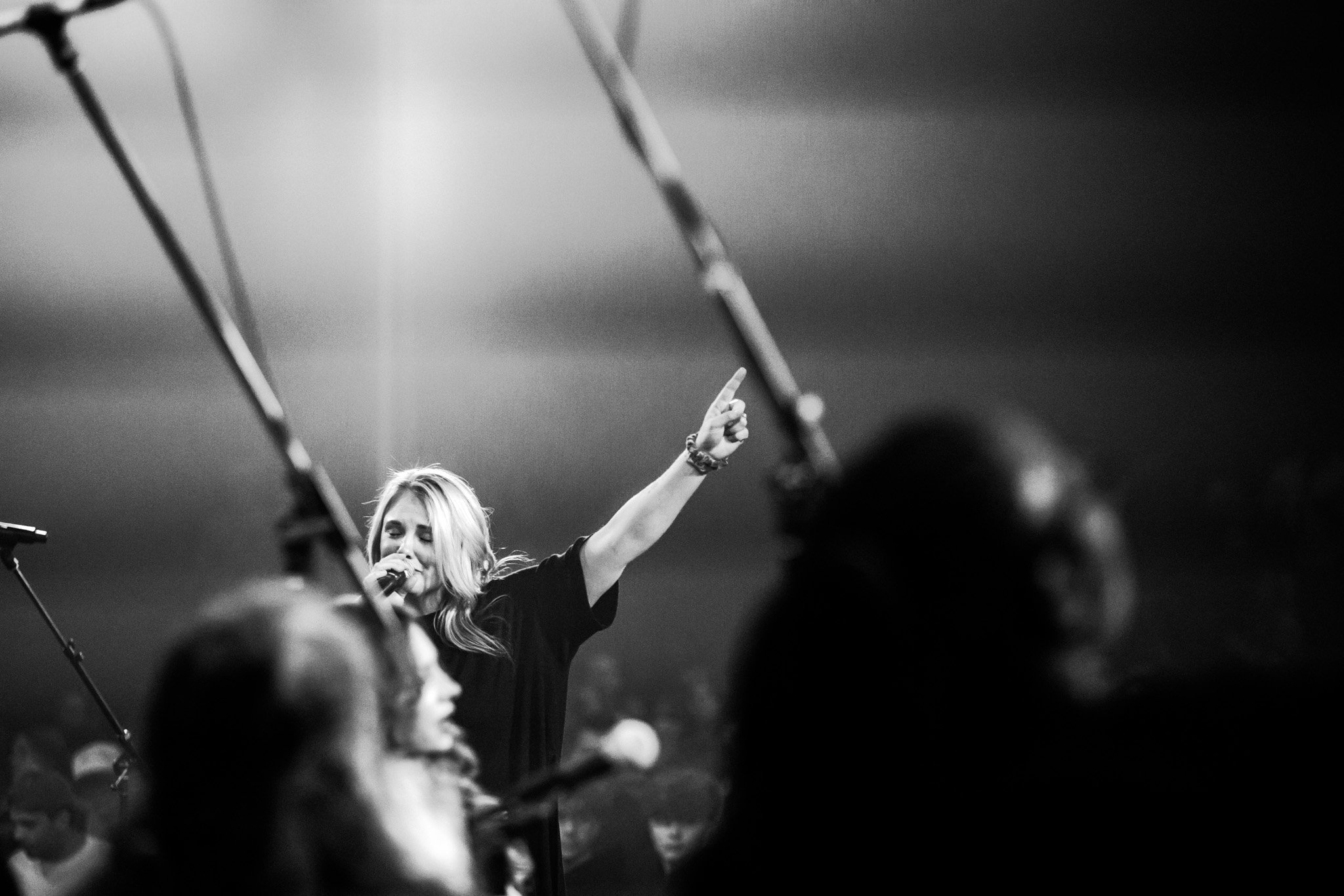 A young woman with blonde hair singing into a microphone on stage, raising her right arm with her index finger pointing upward, surrounded by an audience.