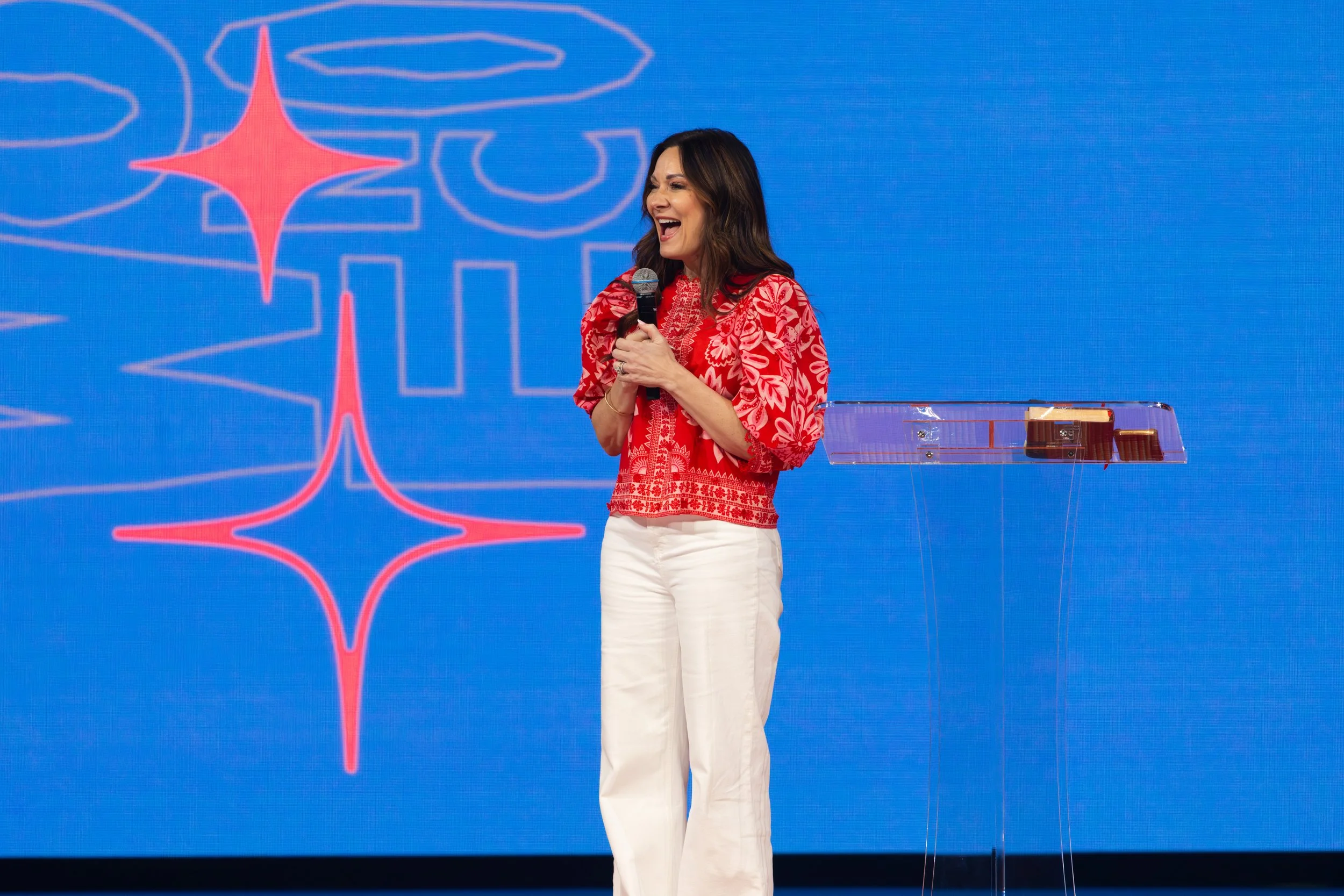 A woman in a red and white patterned blouse and white pants standing on stage, holding a microphone, smiling. Behind her, a large blue screen with a red and white outline of the Pittsburgh Steelers logo.