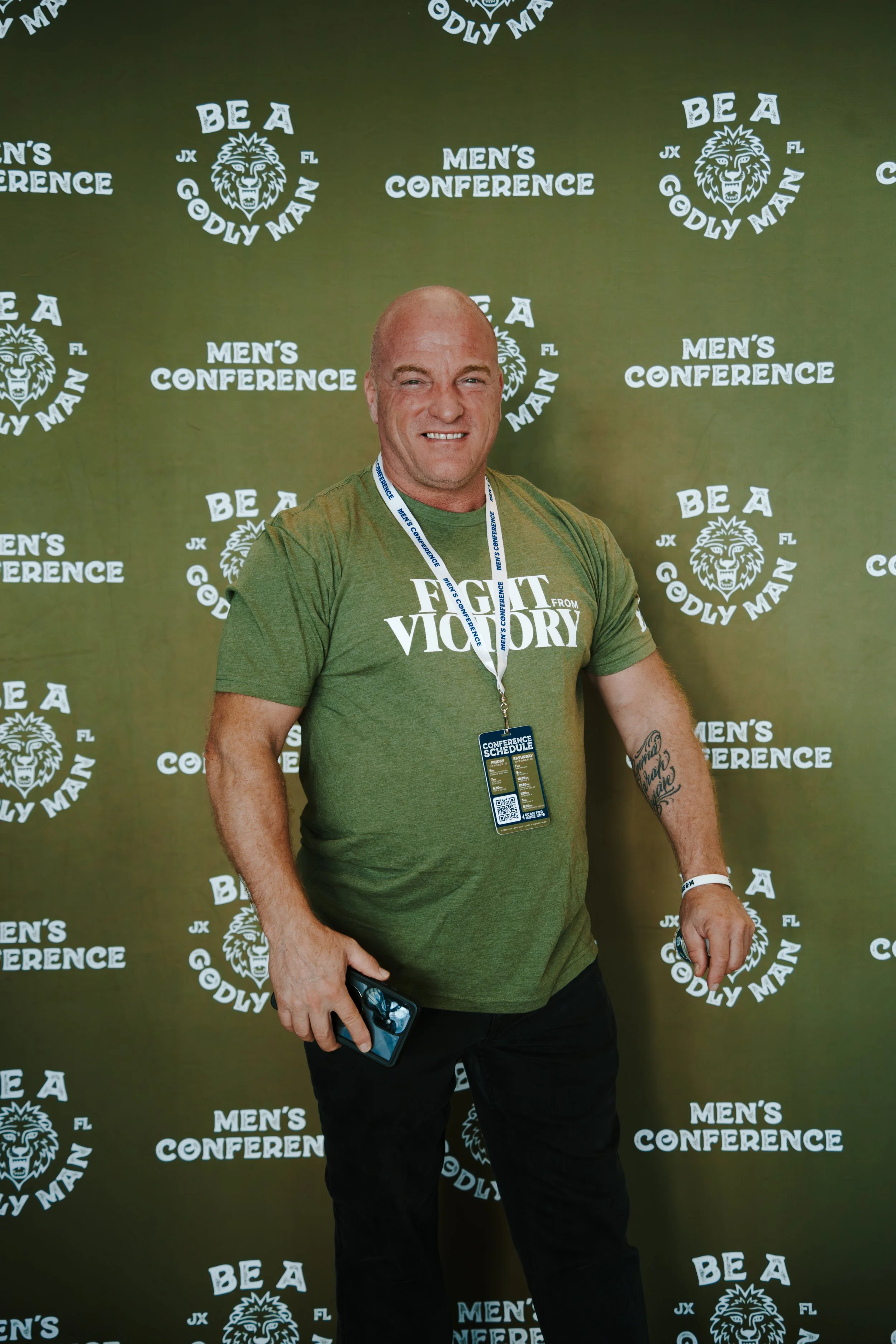 A smiling man in a green t-shirt with the words 'Fight for Victory' standing in front of a green conference backdrop with slogans and a lion logo, holding a conference schedule and a smartphone.