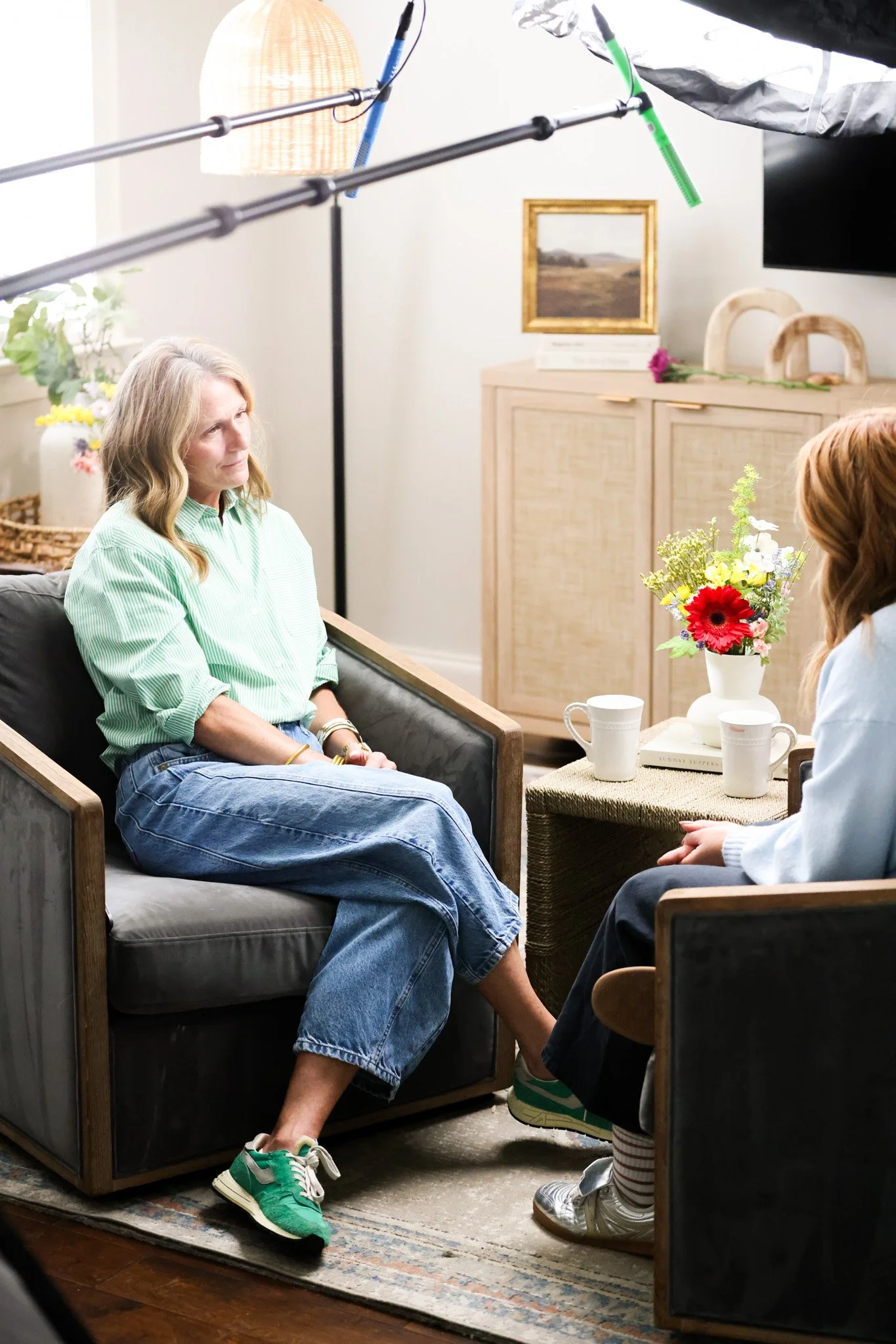 A woman in a green striped shirt and jeans sitting on a sofa, being interviewed in a cozy room with a coffee table, flowers, and artwork on the wall. The interview appears to be recorded with professional lighting equipment.