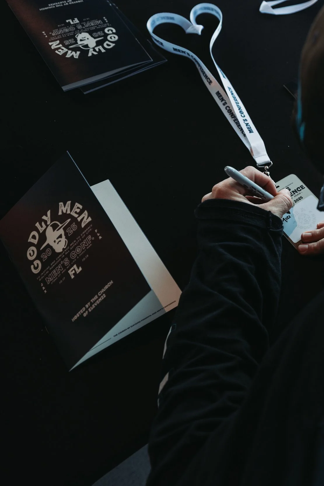 Close-up of a person's hand signing a conference badge at the Goldy Men Men's Conference, with event brochures and a lanyard on a black table.