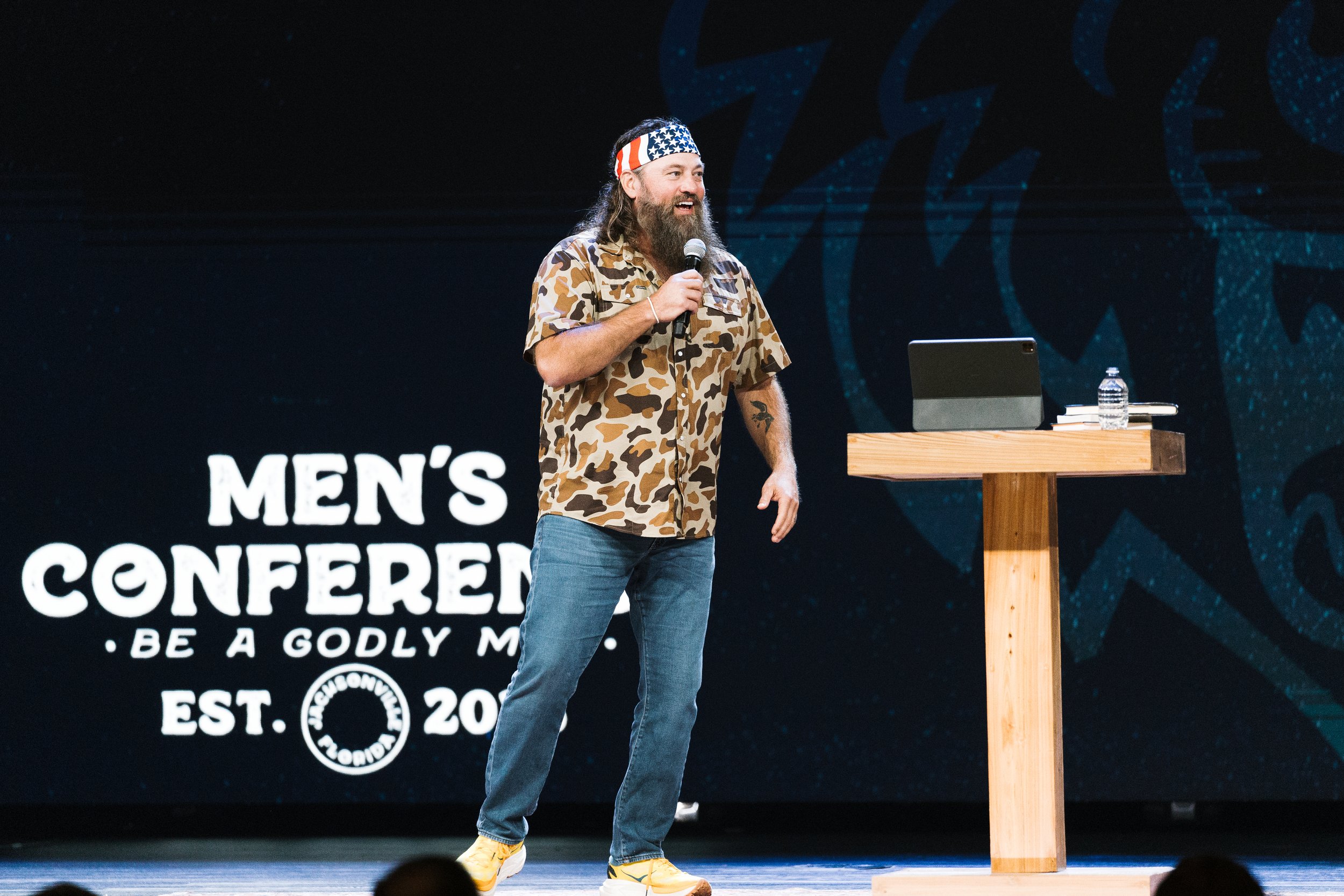 A man with a beard, wearing a camouflage shirt and a headband with American flag design, is speaking into a microphone on stage. There is a wooden podium with a water bottle and papers on it, and a large backdrop with text related to a men's conference.