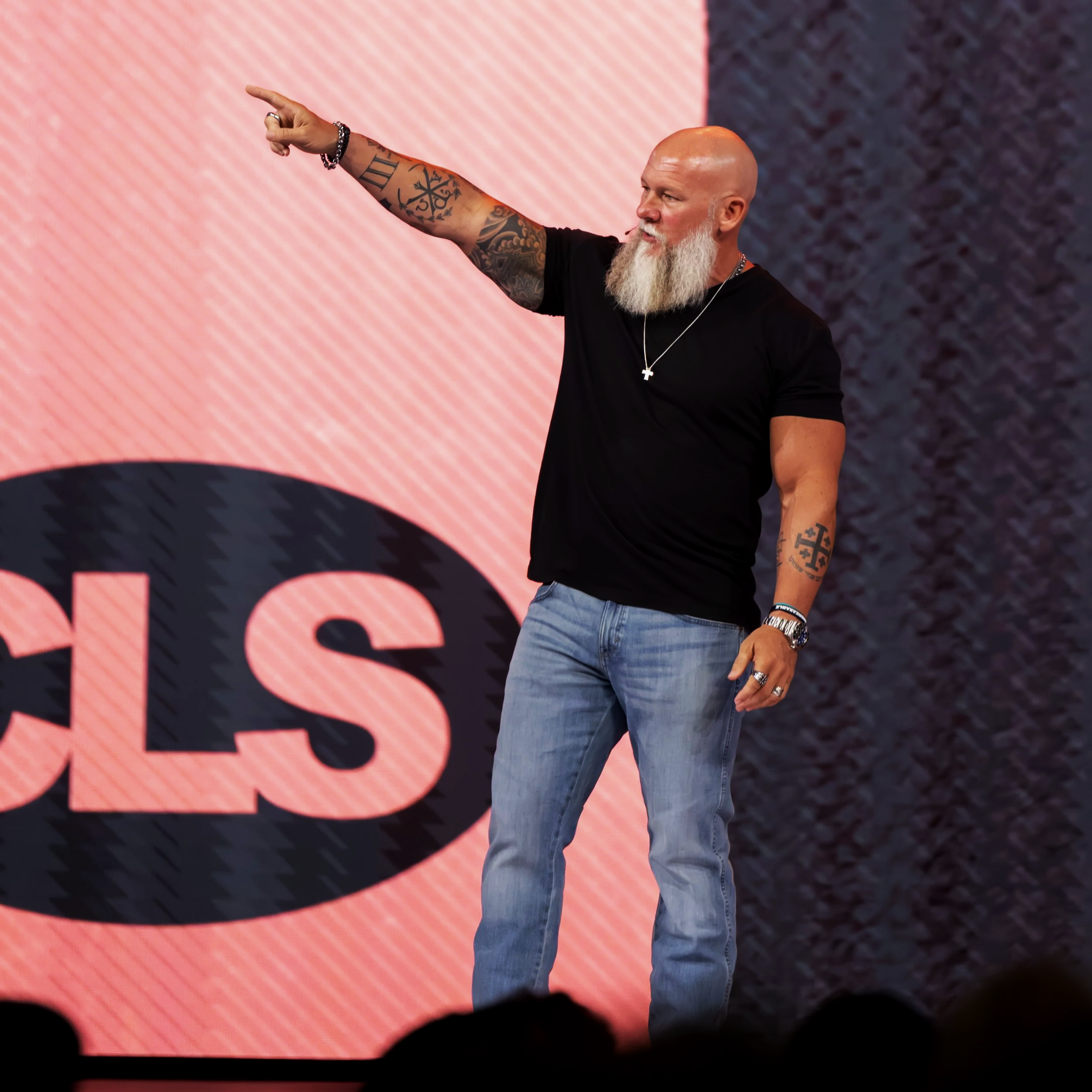 Bald man with a long beard wearing a black t-shirt, gold watch, rings, and a cross necklace, pointing with his left arm towards the left side of the image on a stage with a large pink and dark textured background.