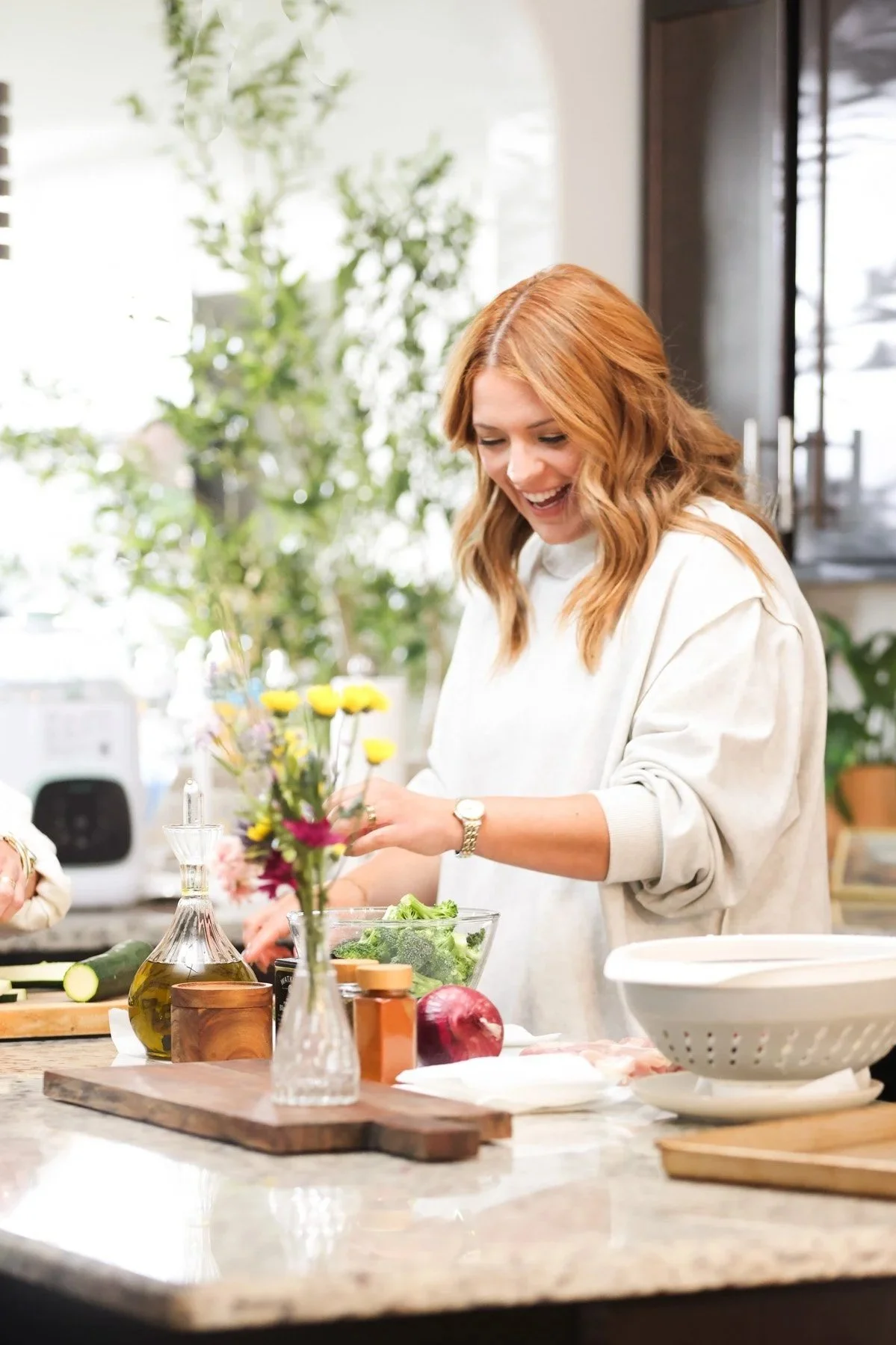 A woman with red hair and a white sweater preparing food in a bright kitchen with a marble countertop, surrounded by cooking ingredients, utensils, and a vase with flowers.