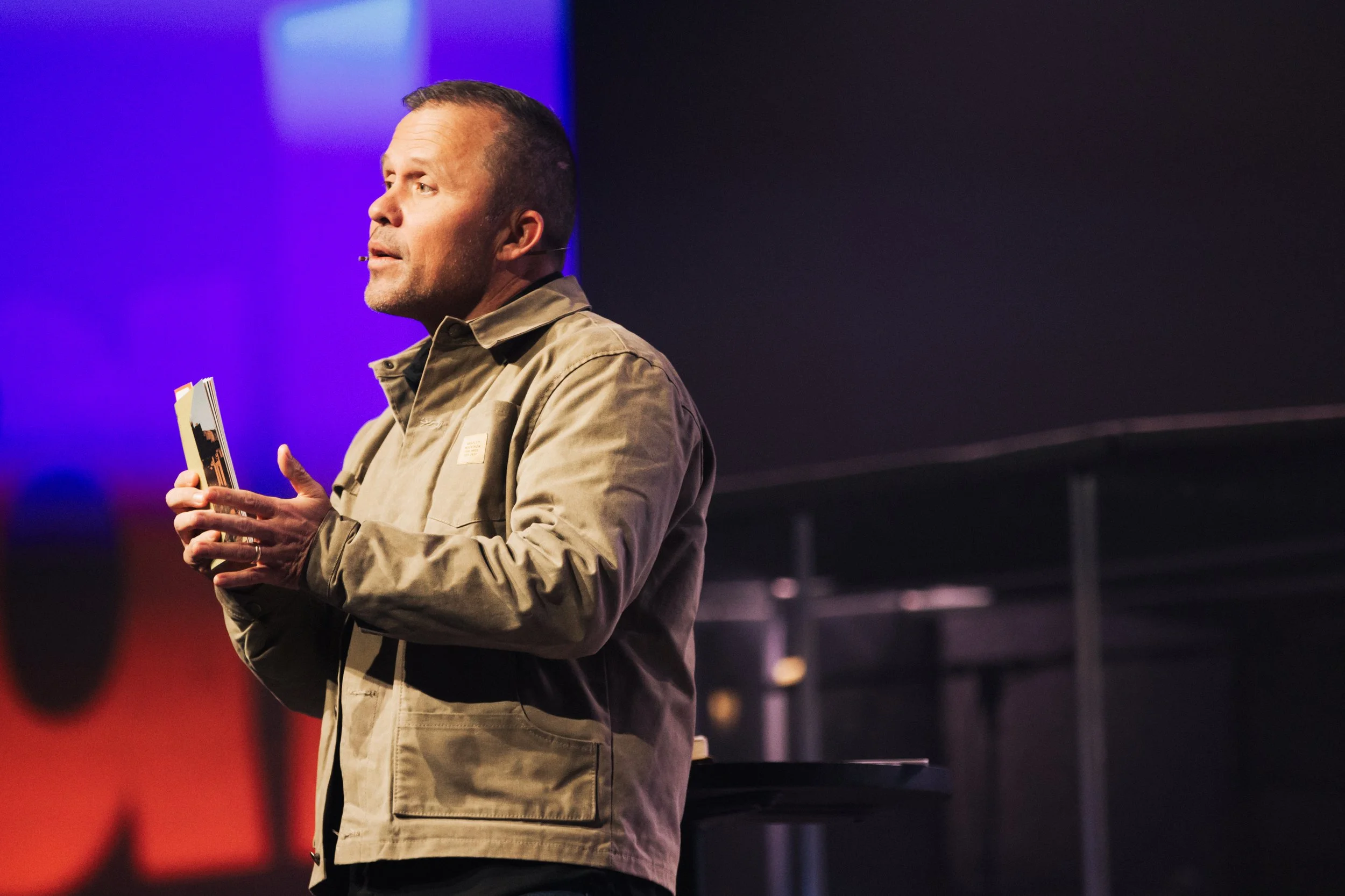 A man with short dark hair and a stubble beard, wearing a tan jacket, is holding a book and presenting on stage against a backdrop with blue and purple lighting.