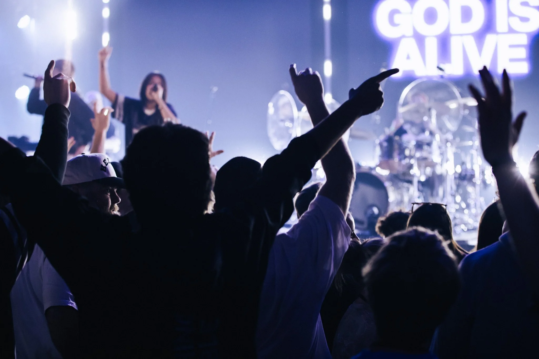 A crowd of people at a concert or worship event with hands raised. The stage has a band and a large screen displaying the text 'GOD IS LOVE' in the background.