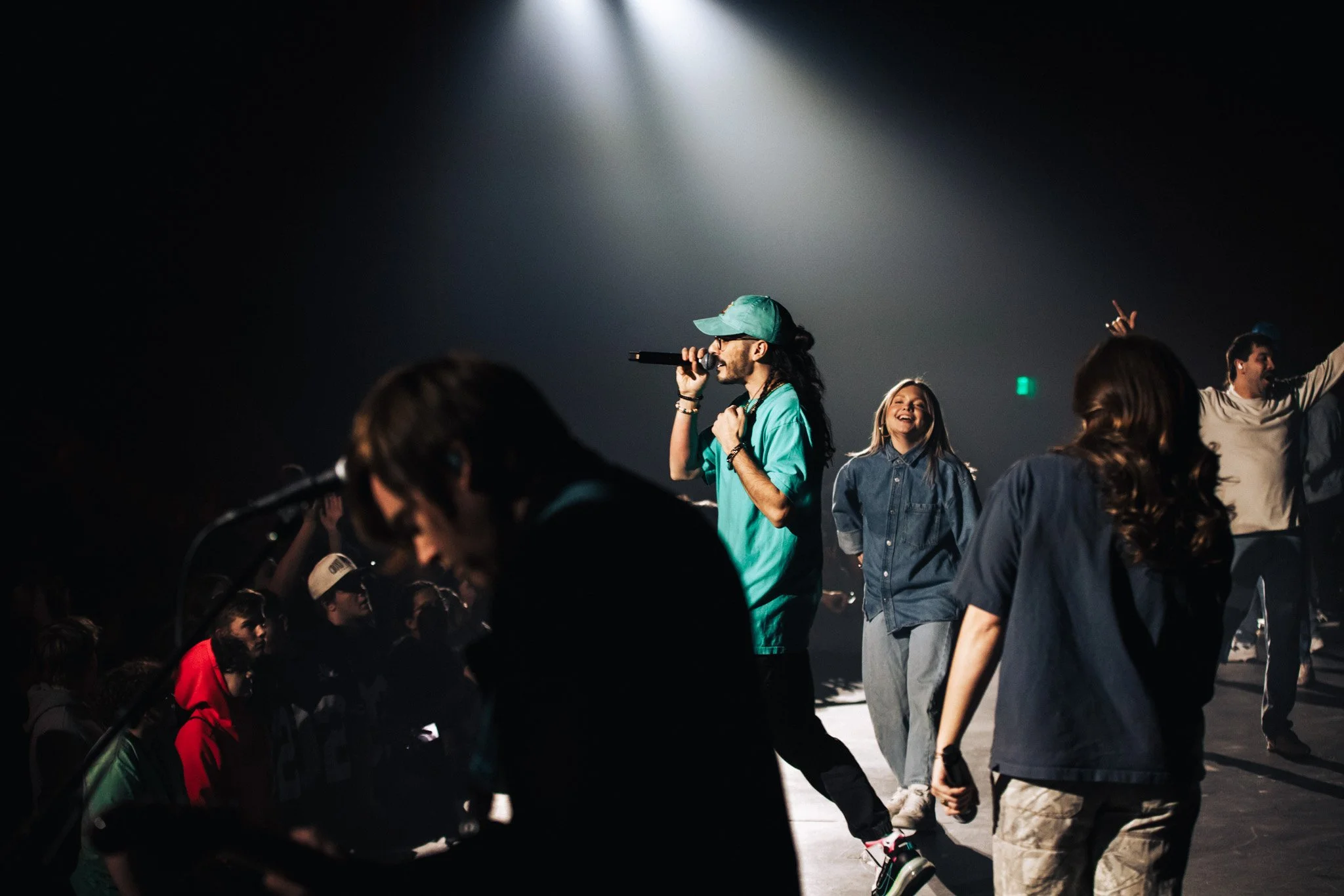 People gathered in a dimly lit space with a spotlight shining down, a woman holding a microphone on stage, surrounded by others, some dancing or singing along.
