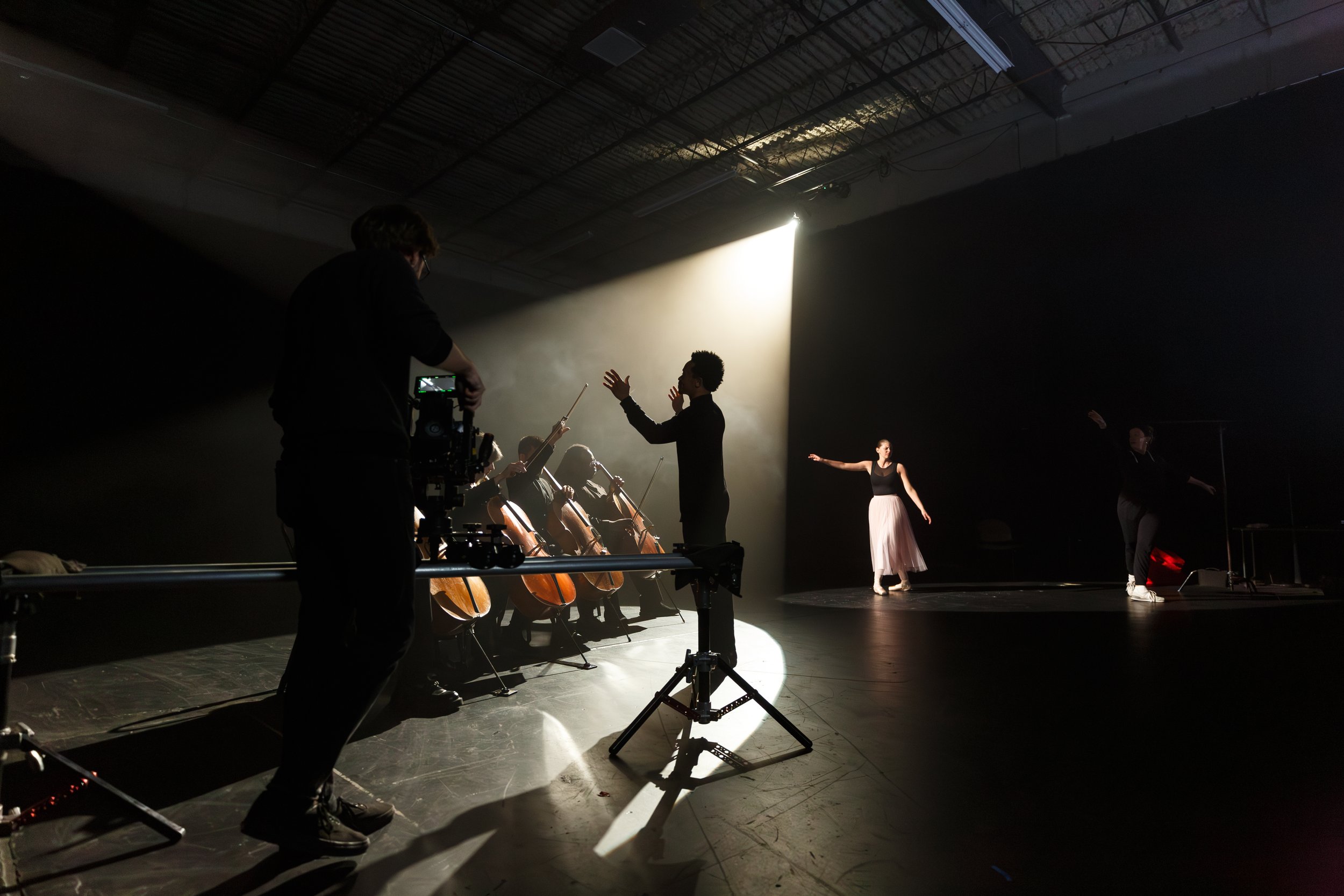 A ballet dancer in a black top and pink tutu practicing on stage with a conductor and musicians in the background, illuminated by a spotlight in a dark rehearsal space.
