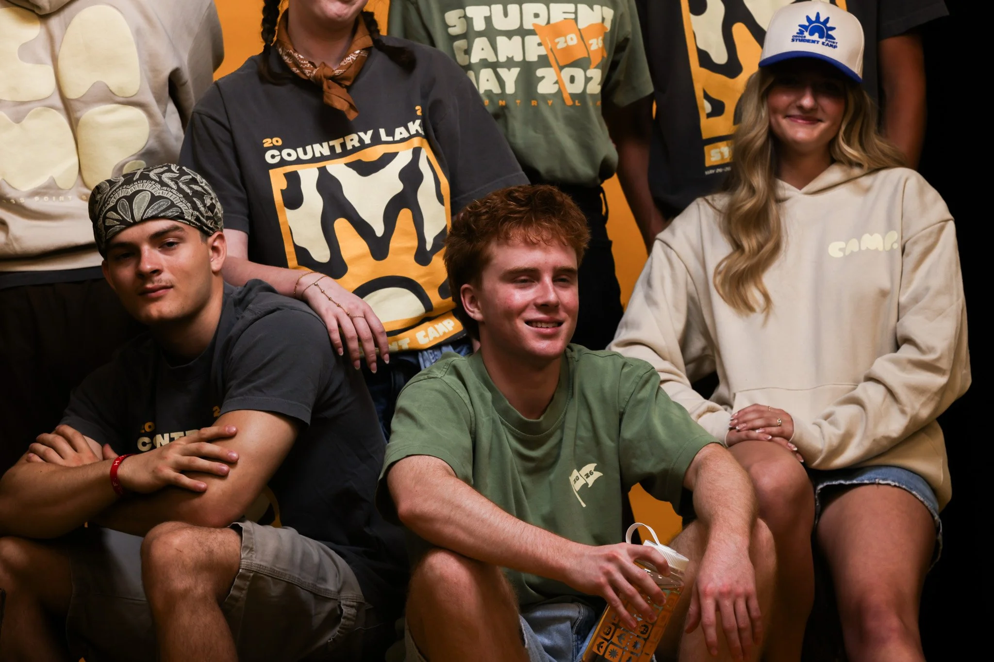 Group of smiling young people wearing outdoor or camp-themed clothing, including t-shirts with 'Country Lake' and 'Student Camp' logos, posing against an orange backdrop. One girl has long blonde hair and is wearing a white cap and hoodie, another person has braided hair and wears a bandana. The group appears to be at a camp or outdoor event.