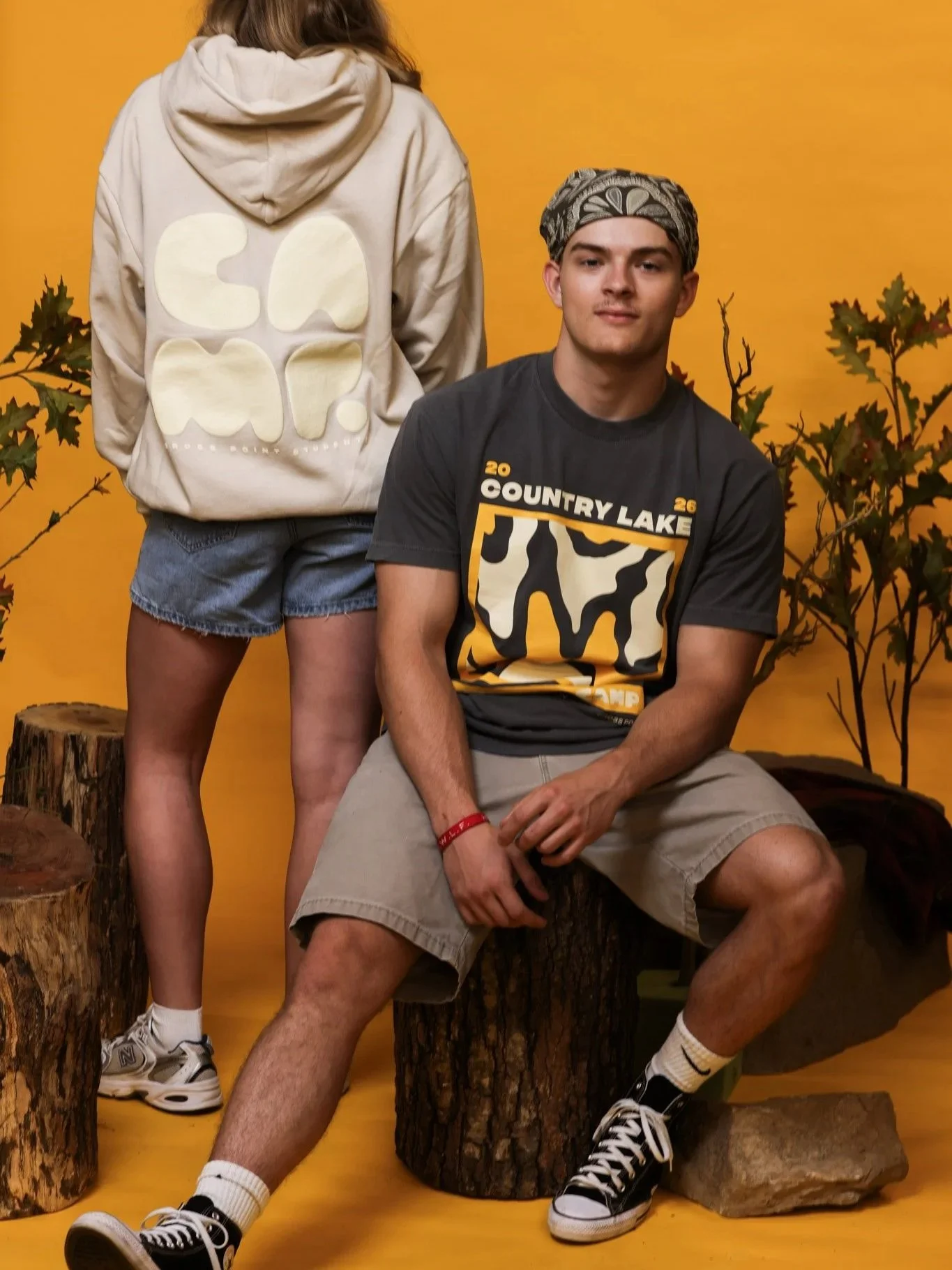 A young man wearing a bandana, a graphic t-shirt, and shorts, sitting on a tree stump, with a woman's back visible standing next to him. The woman wears a hoodie with a design on the back and denim shorts, against a yellow background with autumn leaves.