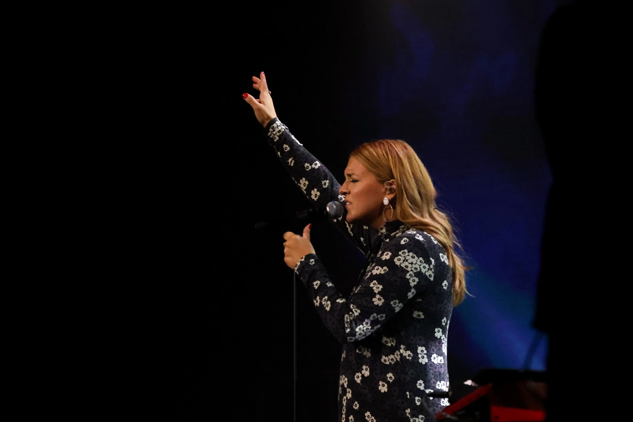 A woman with red hair singing into a microphone while raising her left hand, dressed in a black dress with white floral patterns, against a dark background.