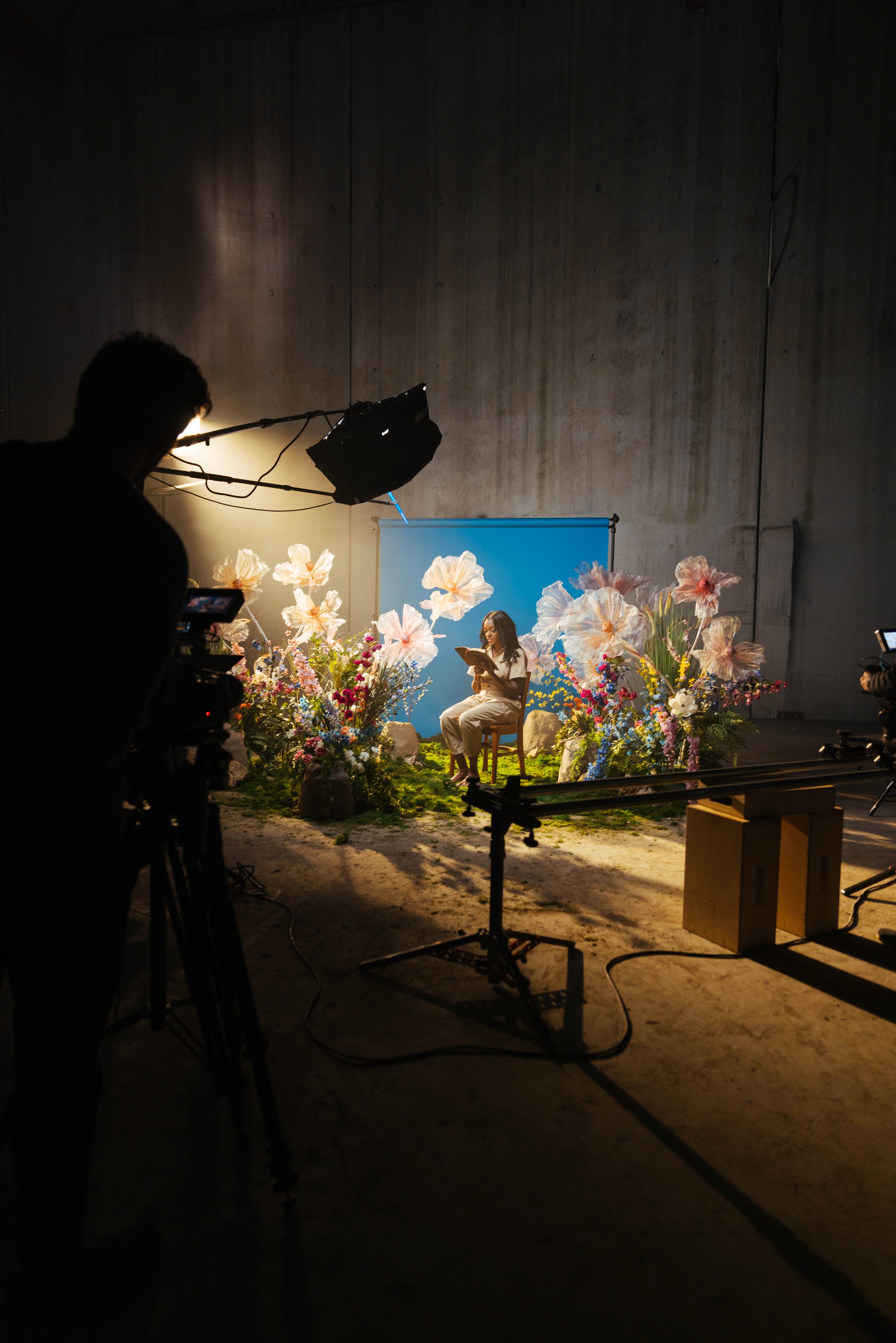 A woman sitting on a chair in front of a blue backdrop, surrounded by large flowers and rocks, with a person behind the camera and studio lights overhead, on a film or photo set.