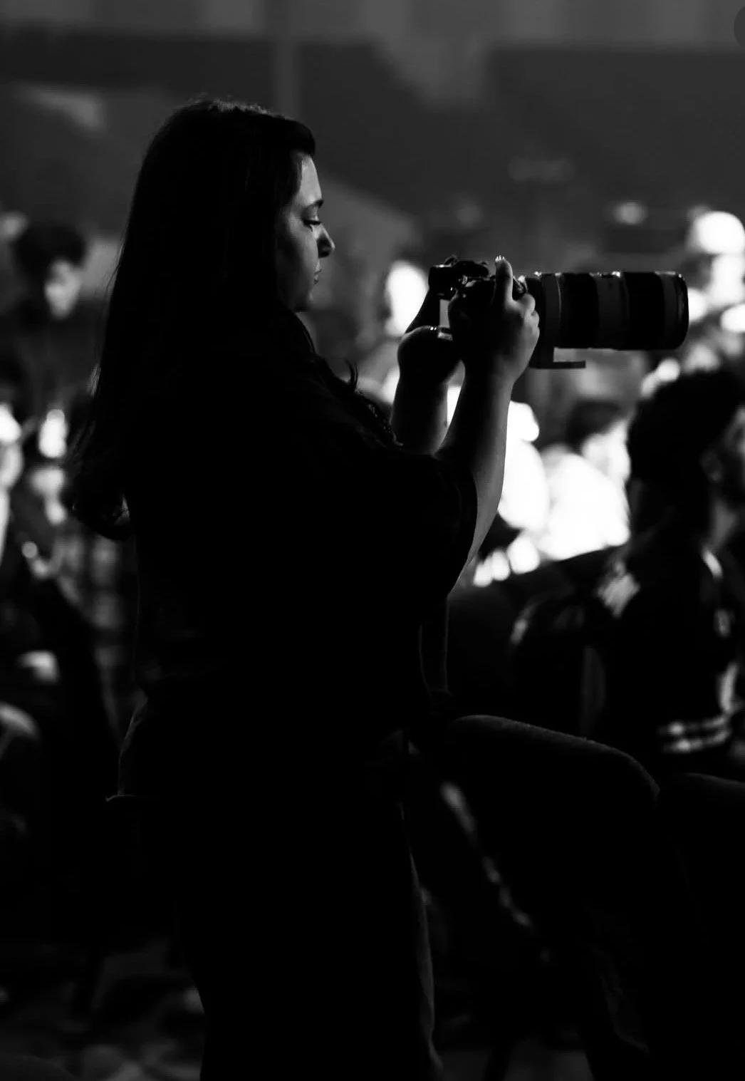 A woman sitting in a dark setting, holding a professional camera with a large telephoto lens, possibly taking photographs at an event or in a studio.